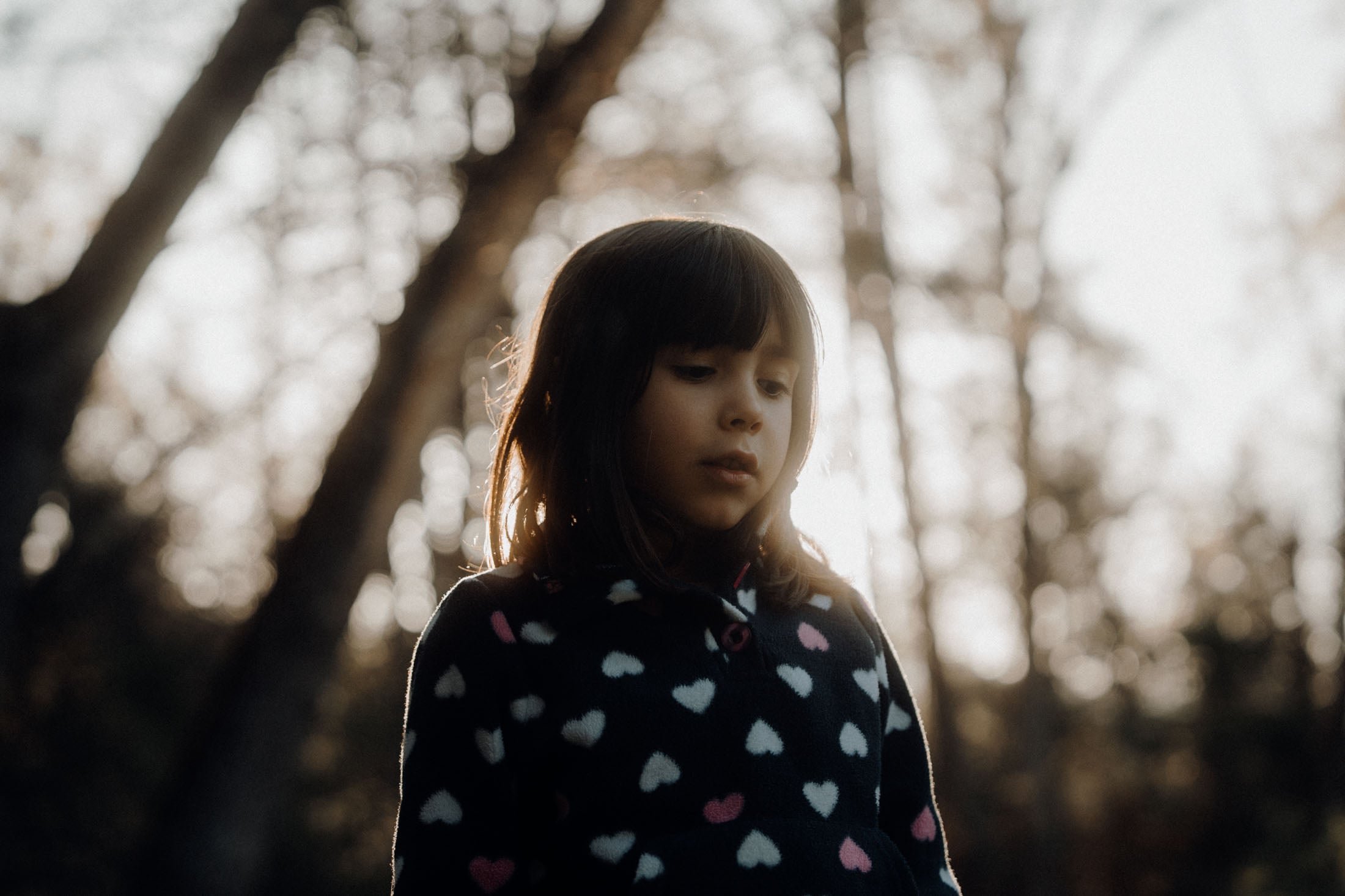 A young girl in a heart-patterned sweater stands in a wooded area, softly illuminated by sunlight filtering through the trees.