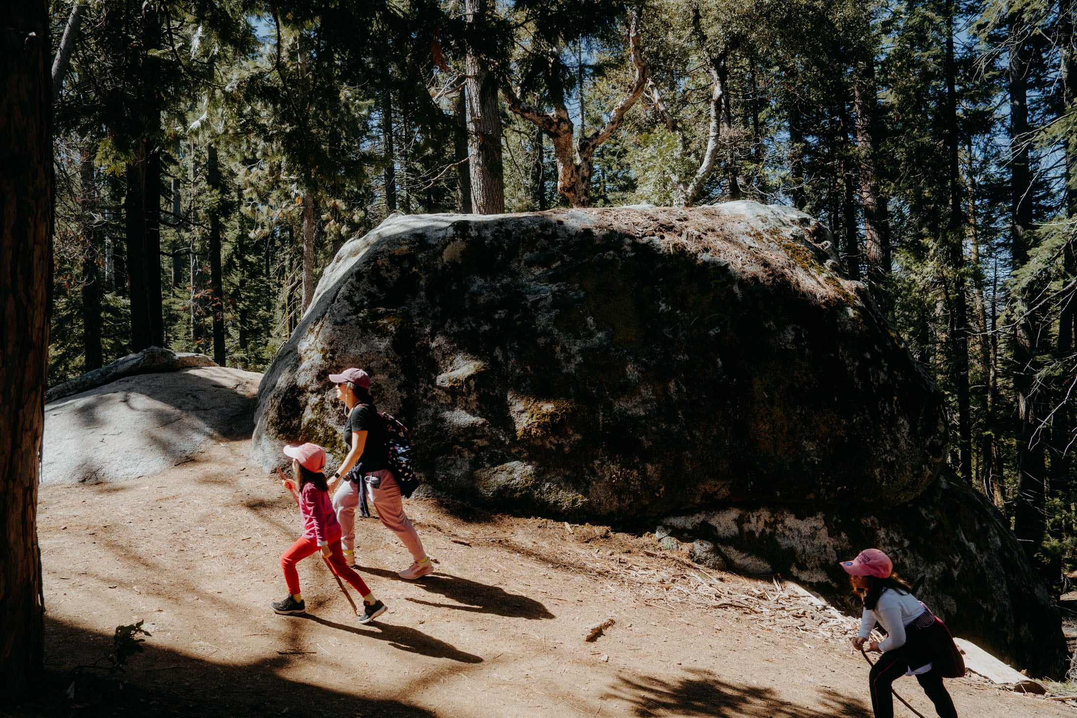 Two children hiking in a forested area, climbing a path next to a large rock formation.