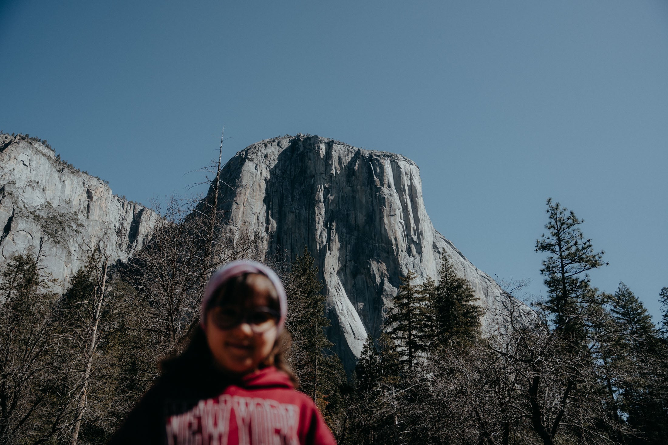A child standing in front of a large rock formation in Yosemite National Park, with trees surrounding the area and a clear blue sky overhead.