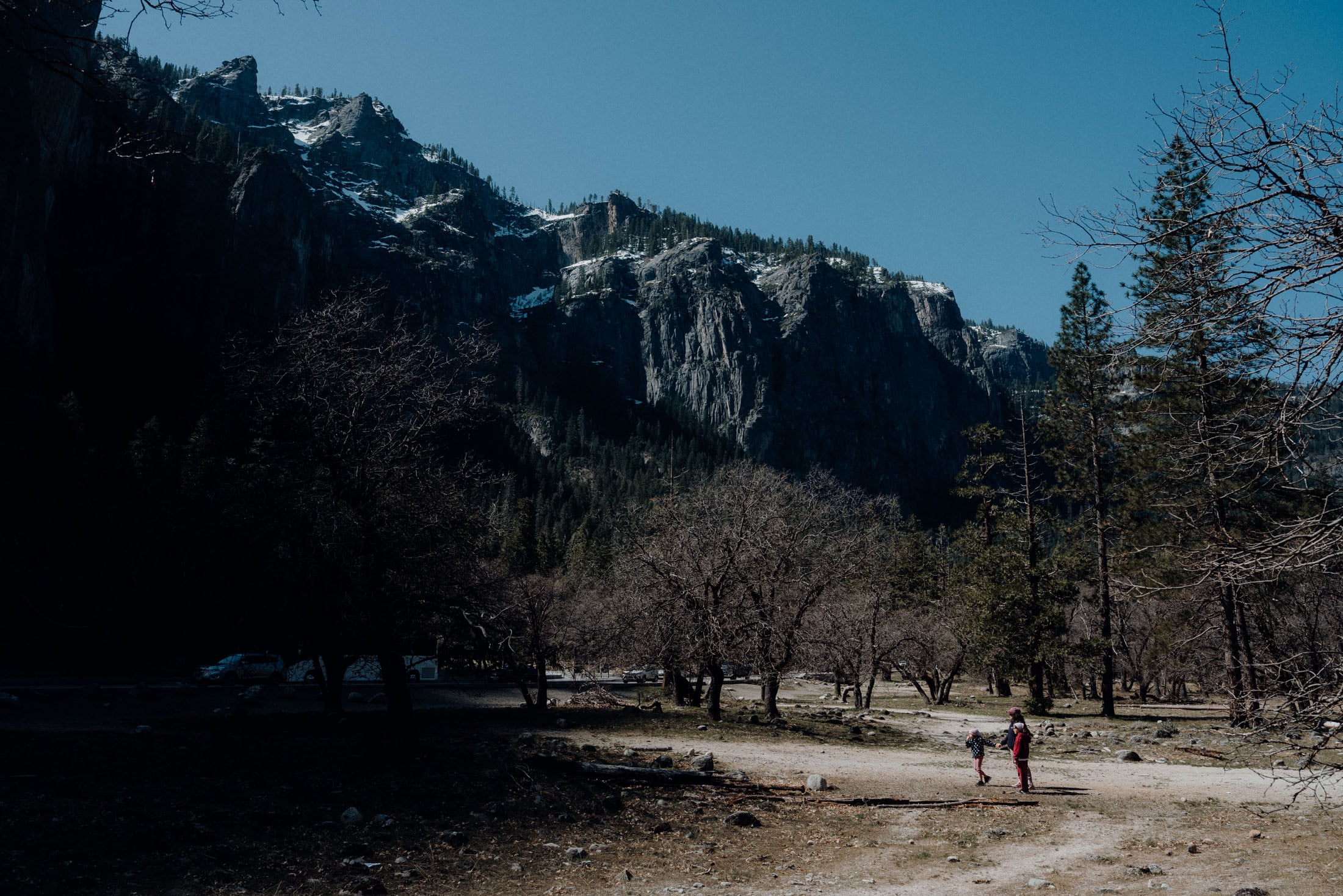 A scenic view of Yosemite National Park featuring towering cliffs and snow-capped mountains, with two people walking along a dirt path surrounded by sparse trees.