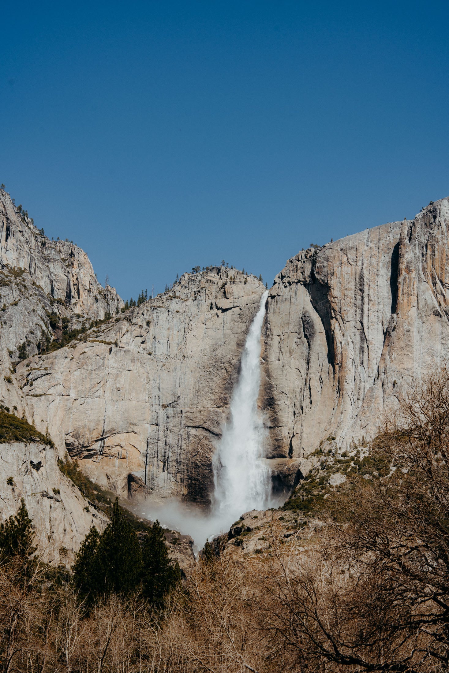 A stunning view of a waterfall cascading down a rocky cliff, surrounded by towering granite formations and a clear blue sky.