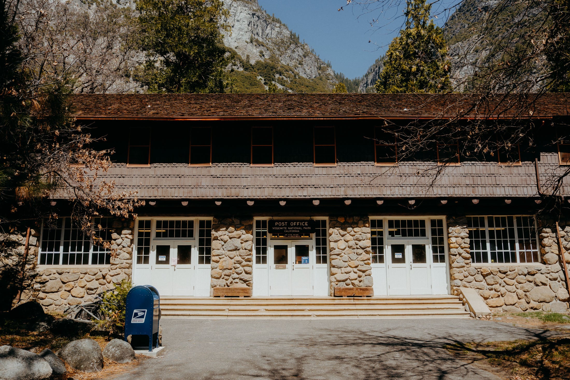 Exterior view of the Yosemite National Park Post Office, featuring a stone facade and white doors, surrounded by trees and mountains.