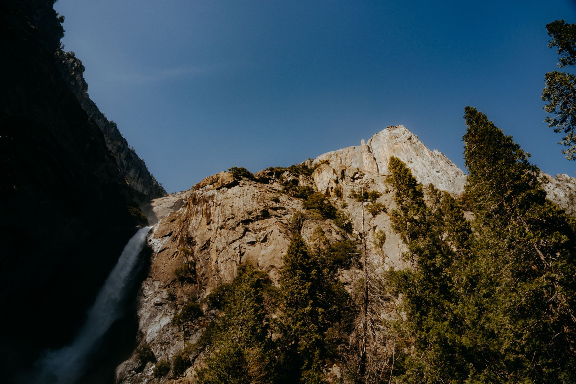 A stunning view of a waterfall cascading down rocky cliffs surrounded by lush green trees, under a bright blue sky.