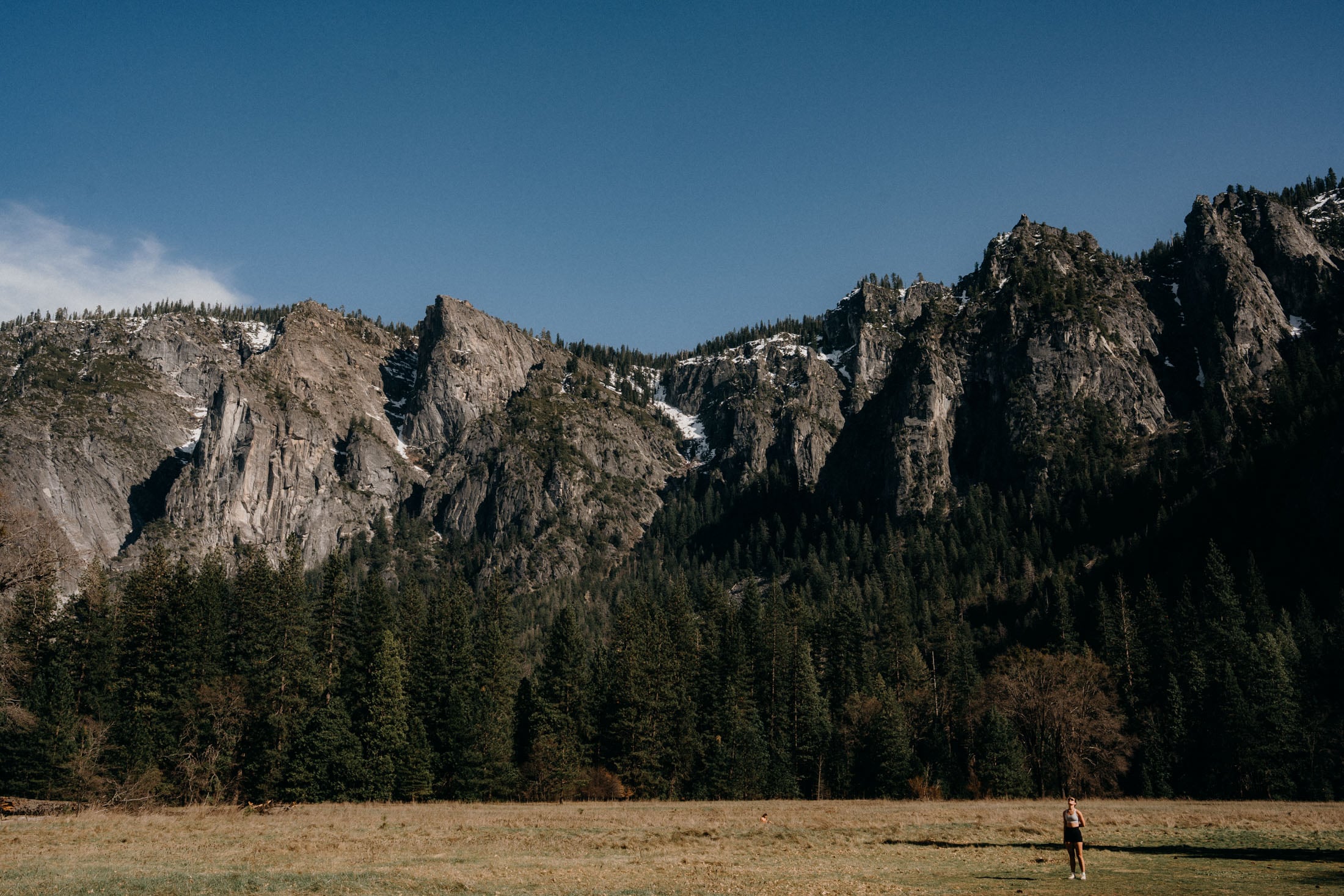 A person standing in a grassy field with towering rocky cliffs and pine trees in the background under a clear blue sky.