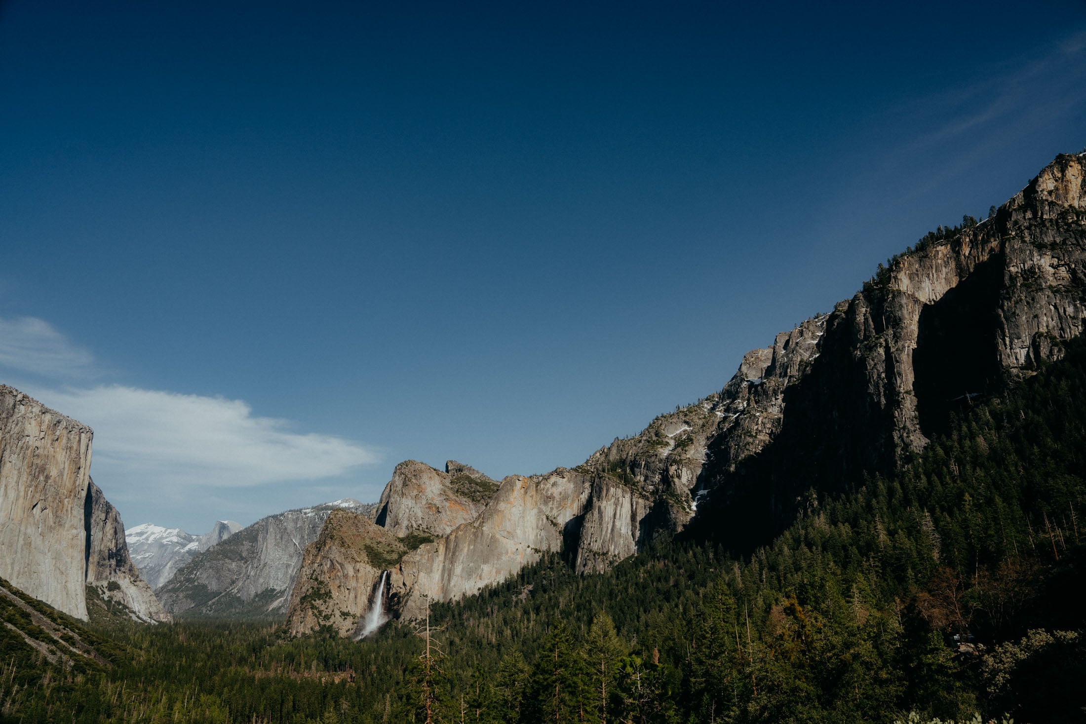 A panoramic view of Yosemite National Park featuring towering granite cliffs, lush green trees, and a clear blue sky.