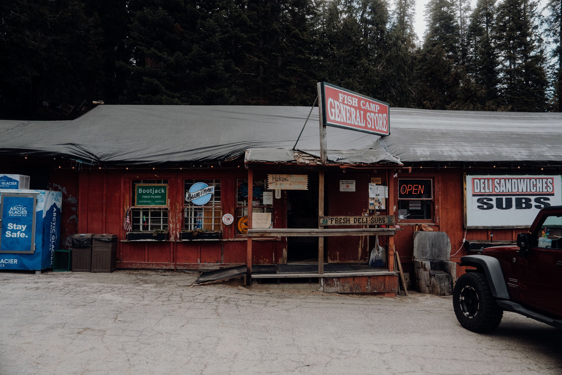 A rustic general store with a red wooden exterior, featuring a sign that reads 'Fish Camp General Store' and an open neon sign. Various signs are displayed on the building, including ones for hiking and deli sandwiches. A black vehicle is parked nearby.