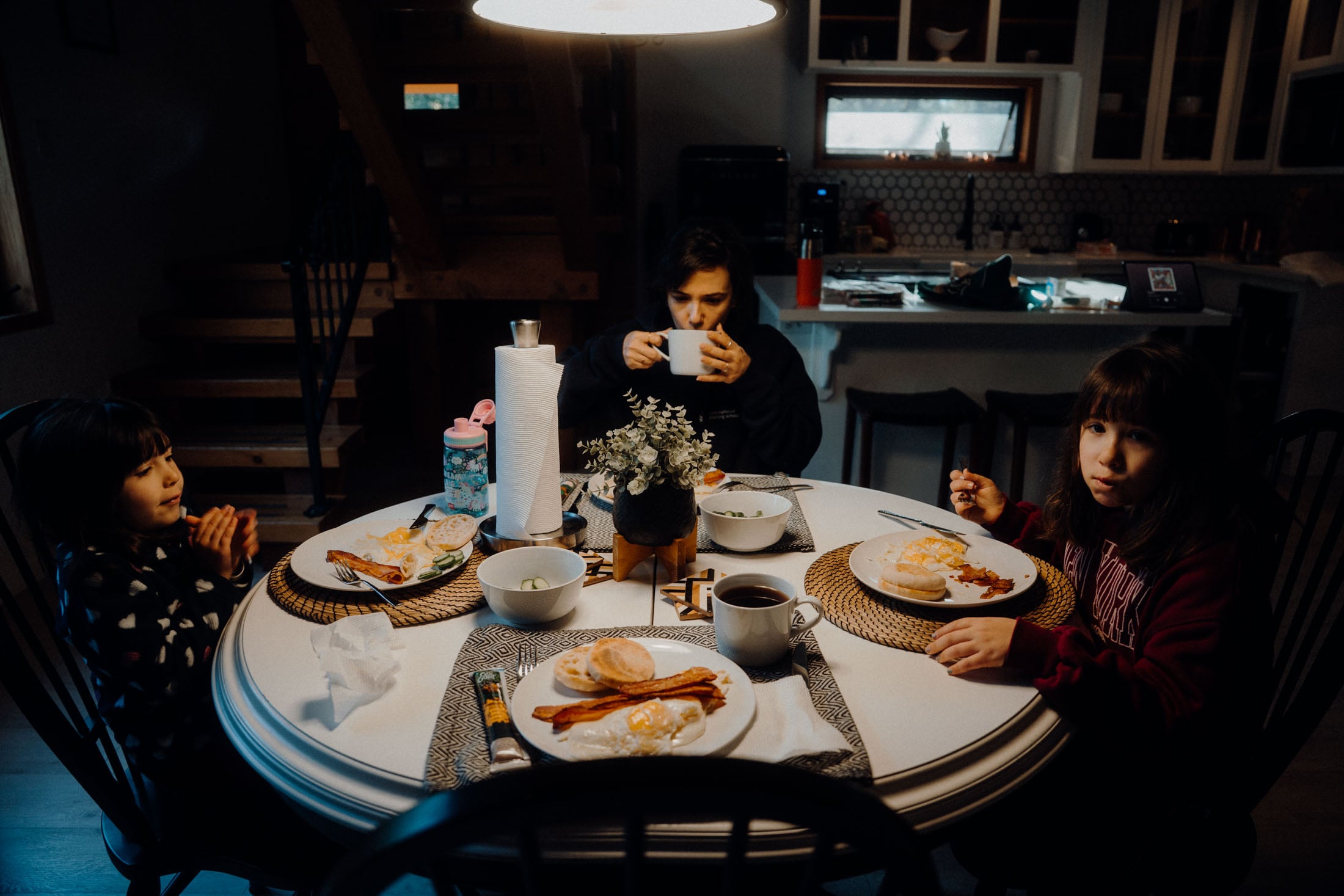 A family enjoying breakfast together at a dining table, with two young girls and a woman sipping from a mug. Plates of food are present, including eggs and bacon, in a cozy, dimly lit kitchen setting.