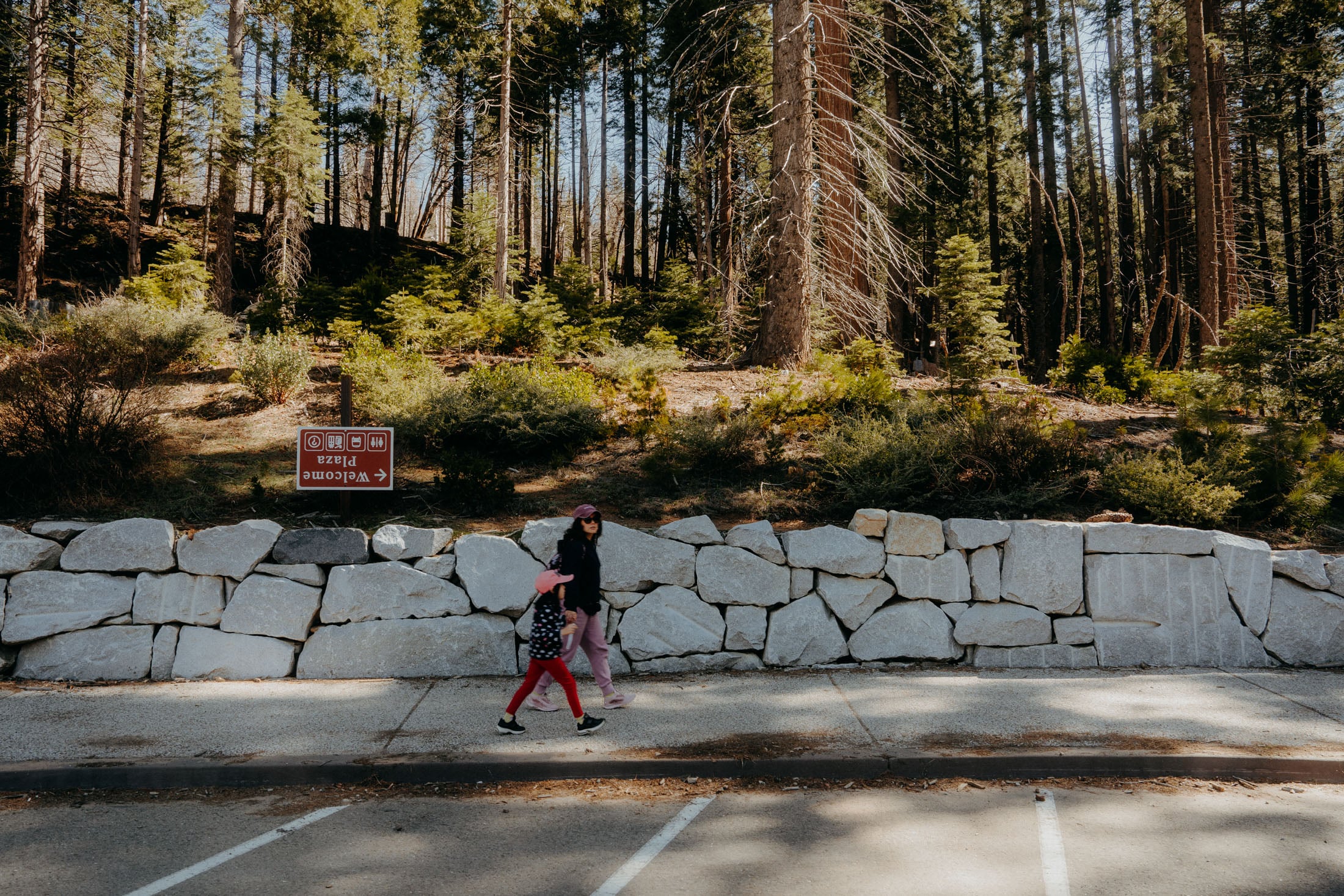 A woman and a child walking along a stone wall in a forested area, with a 'Welcome Plaza' sign visible in the background.