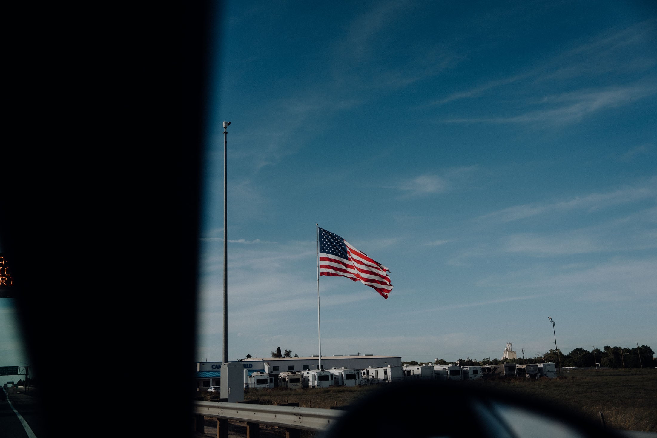 A large American flag waving on a flagpole beside a highway, with blue skies and some clouds in the background.