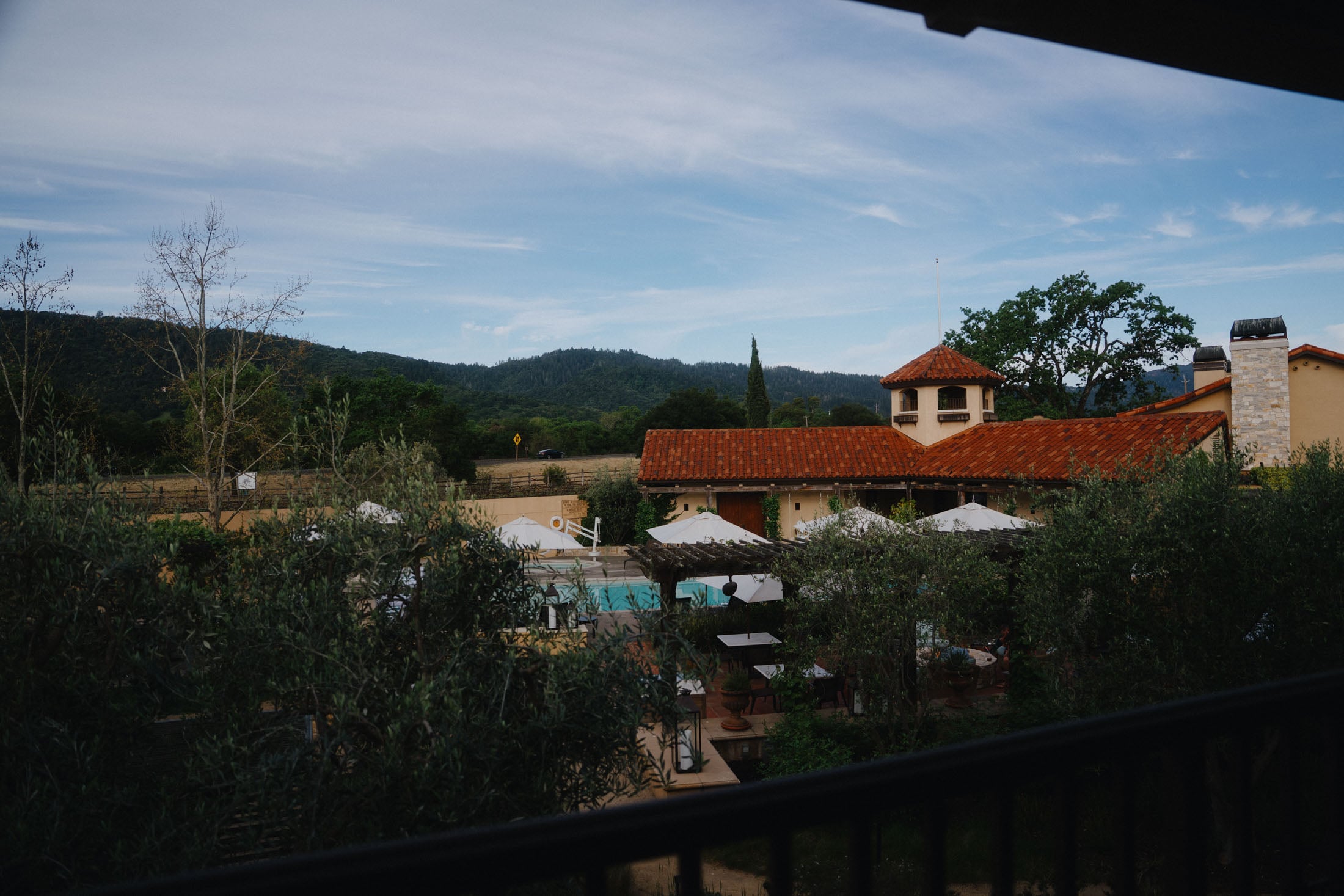 View of a rustic building with a red-tiled roof, surrounded by greenery and a swimming pool, set against a backdrop of rolling hills and blue skies.