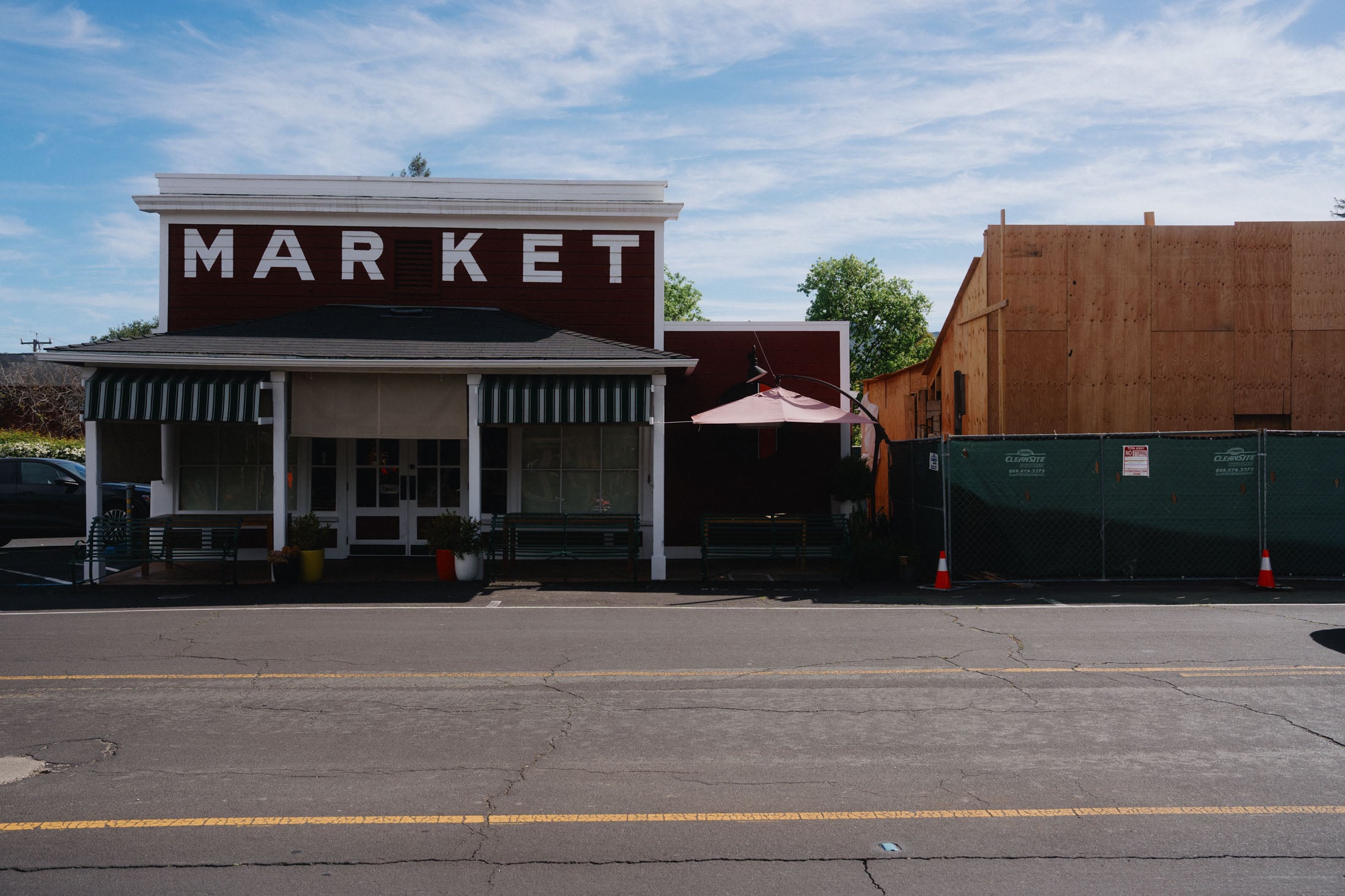 A small market building with the word 'MARKET' prominently displayed, featuring a striped awning and outdoor seating, next to a construction site with wooden walls and fencing.