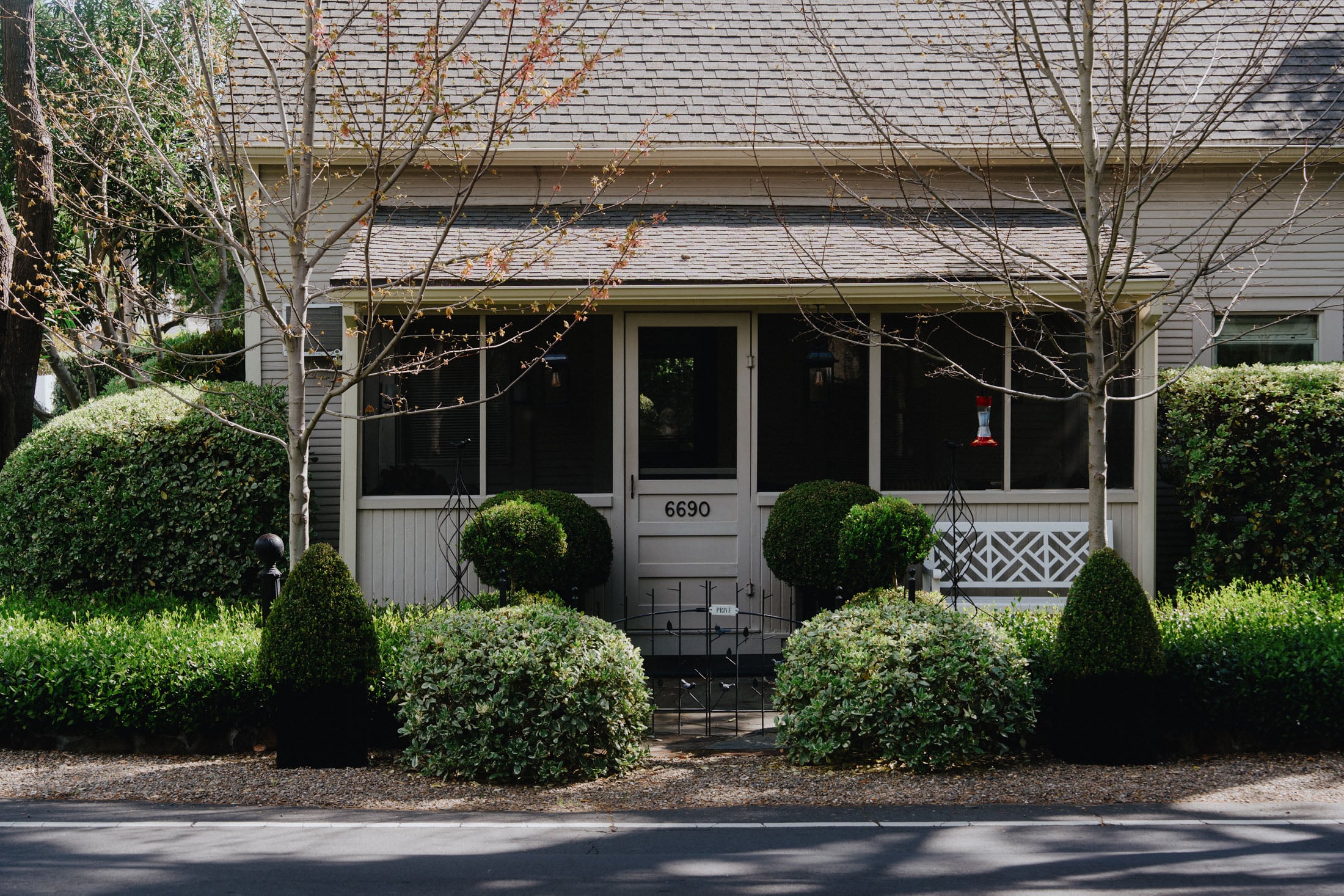 A front view of a house with a porch, featuring a white door labeled '6690', surrounded by manicured bushes and small trees. The yard has green grass and a gravel pathway.