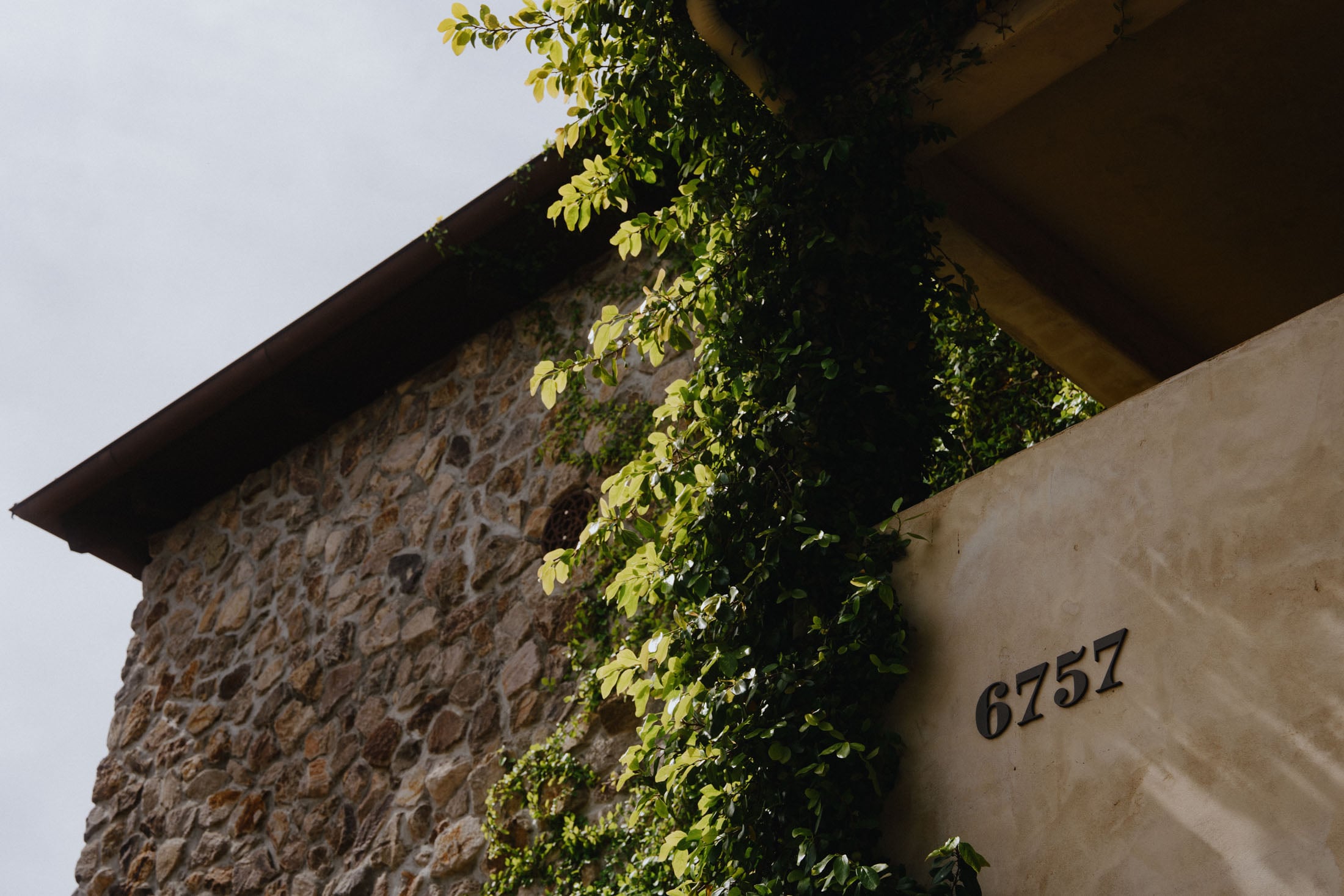 Close-up of a building corner featuring stone and plaster walls, with lush green vines climbing over the surface and a visible house number '6757'.