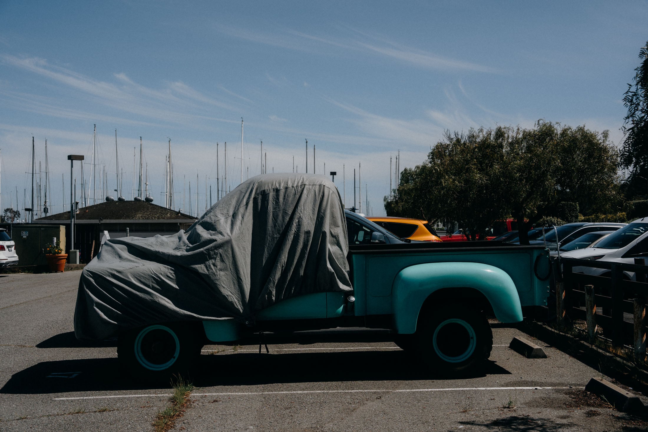 A turquoise pickup truck covered with a gray tarp parked in a lot, with a backdrop of sailboats and a clear blue sky.