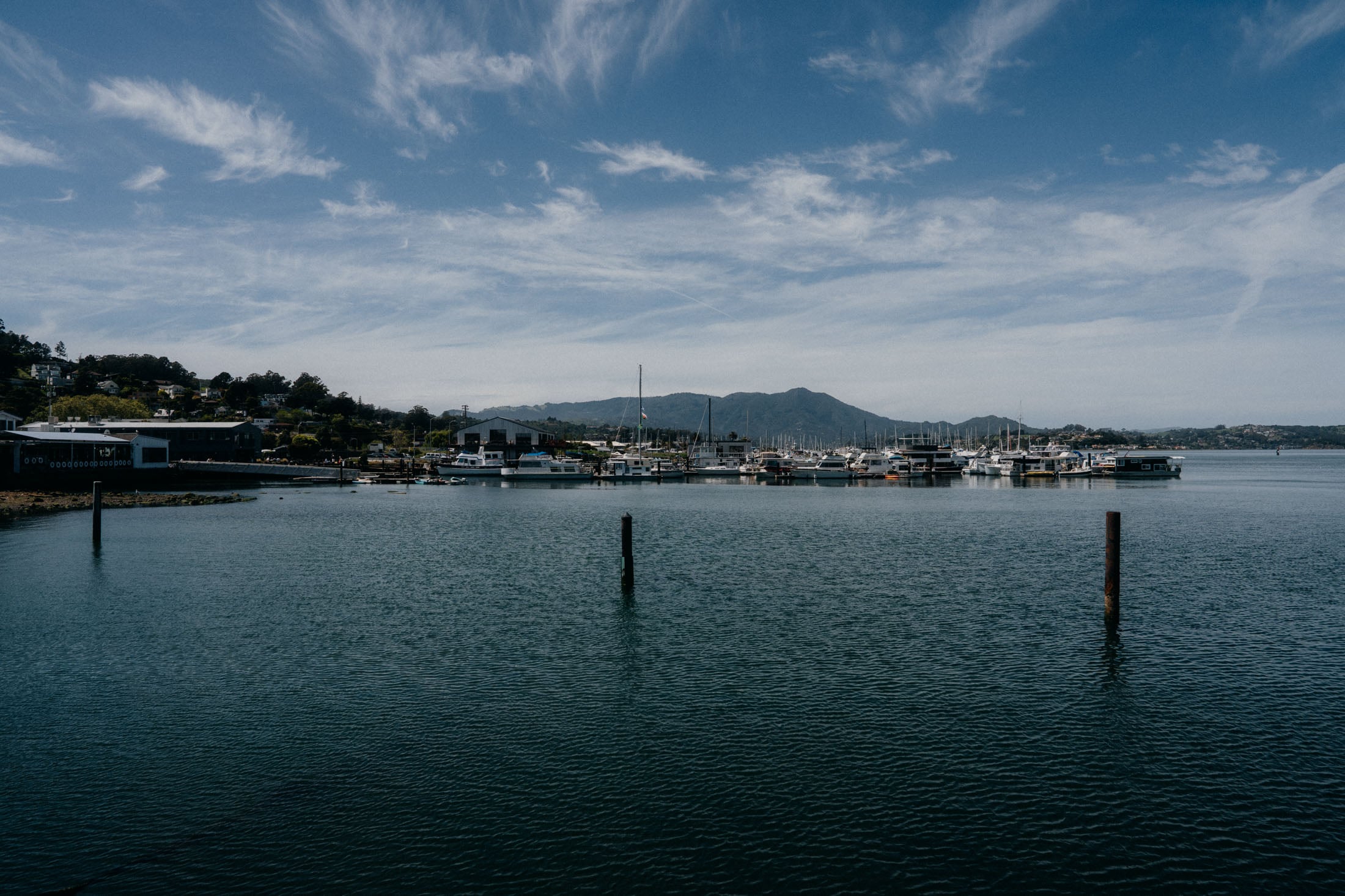 A serene view of a marina with several boats docked, surrounded by hills under a blue sky with scattered clouds.