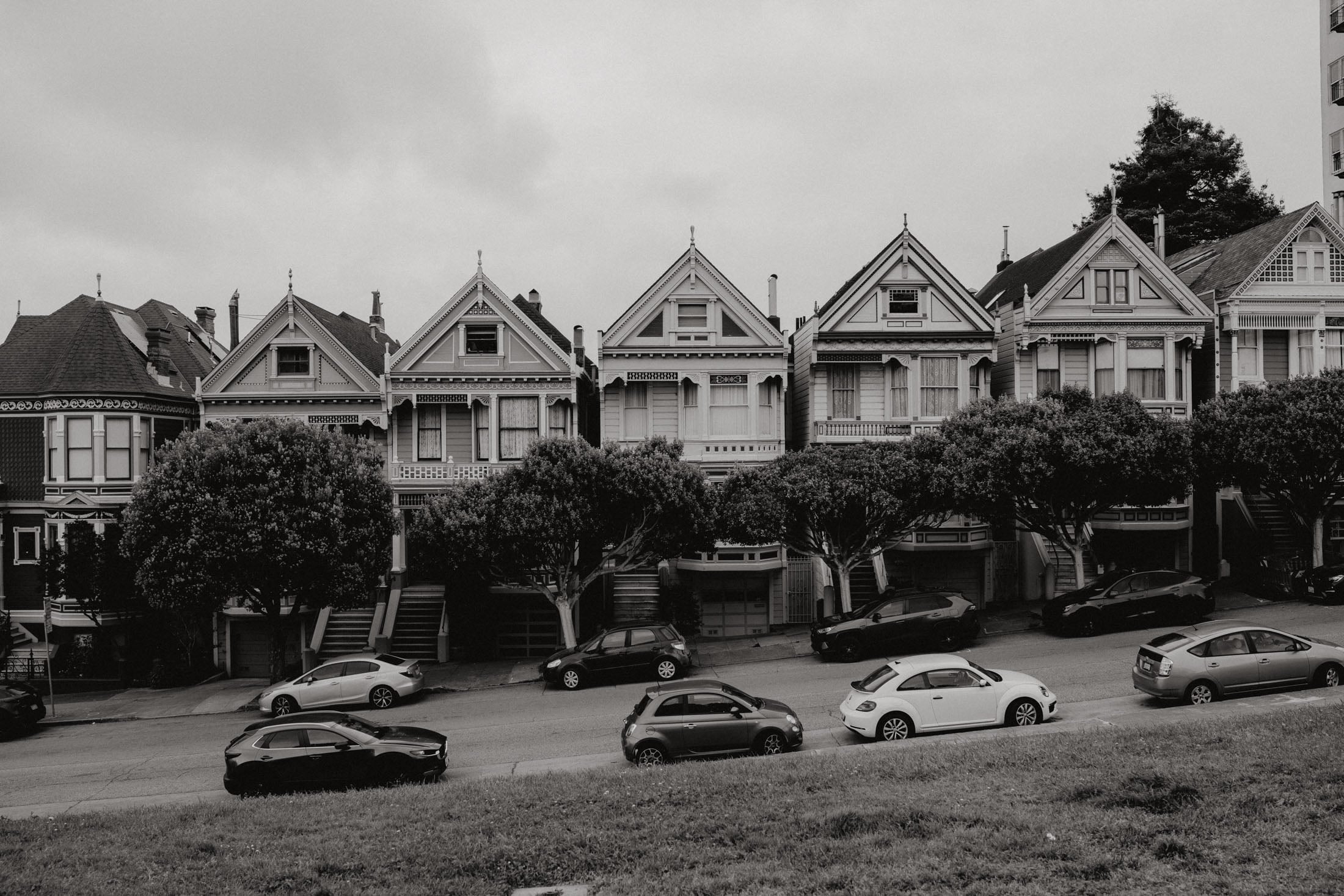 Black and white photograph of Victorian-style houses lined along a street with trees in front and parked cars.