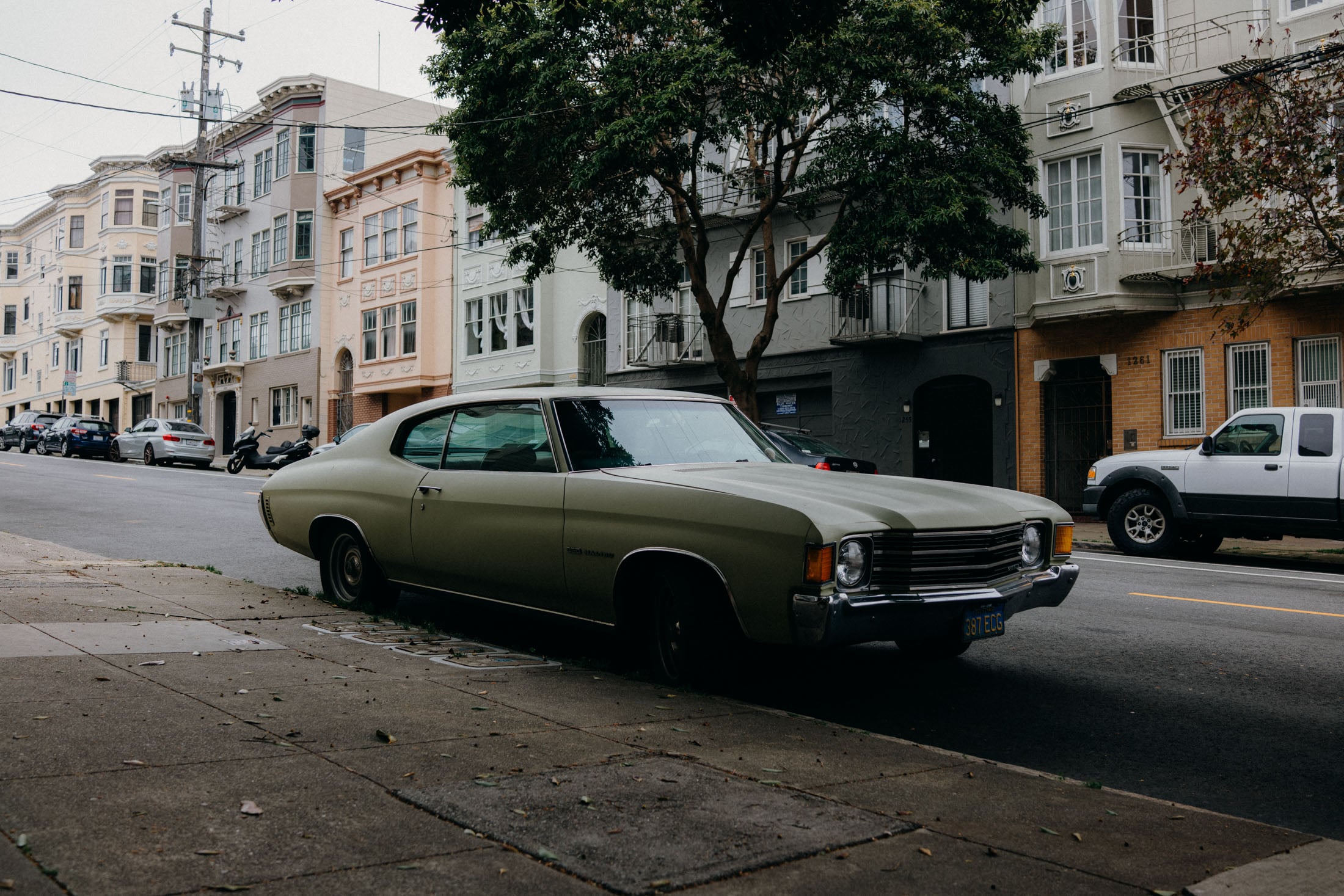 A vintage car parked on the sidewalk in front of colorful residential buildings on a street lined with trees.