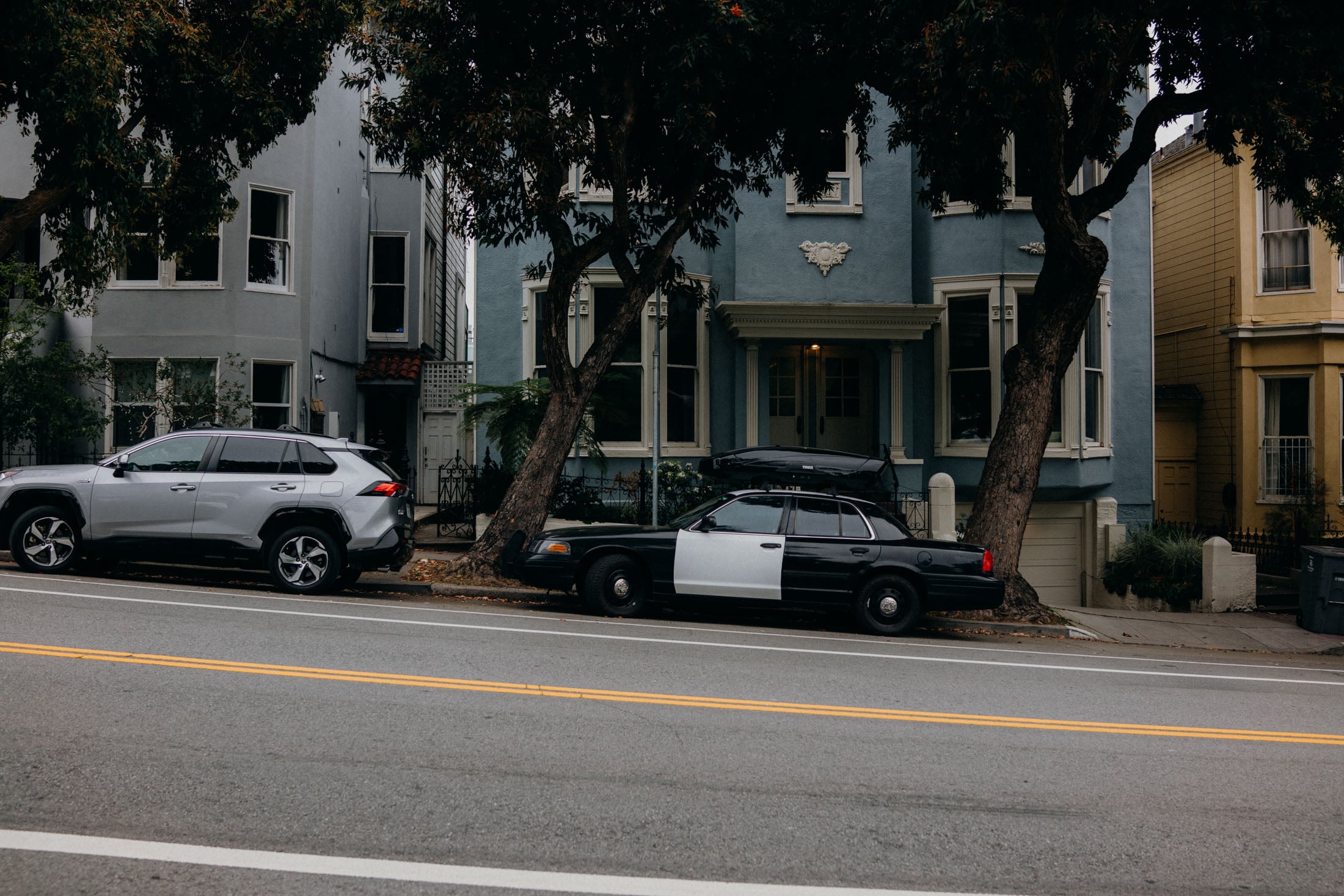 A black and white police car parked on the street next to a silver SUV, with colorful houses in the background.
