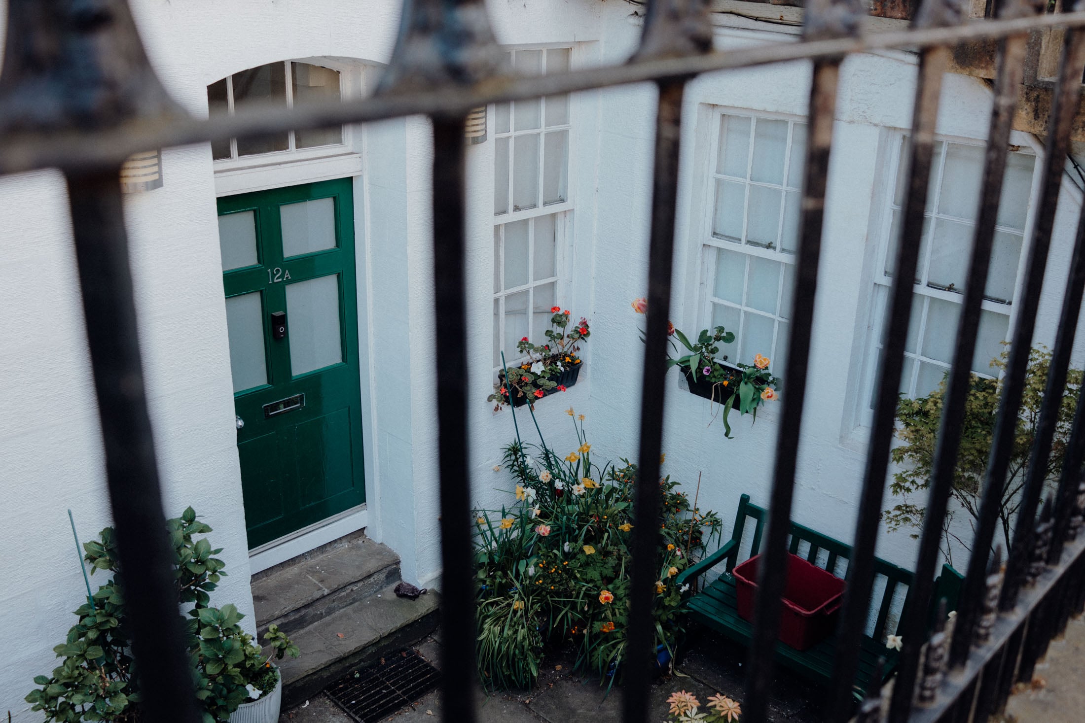 A green door labeled '12A' flanked by two windows with planters filled with flowers, viewed through iron railings.