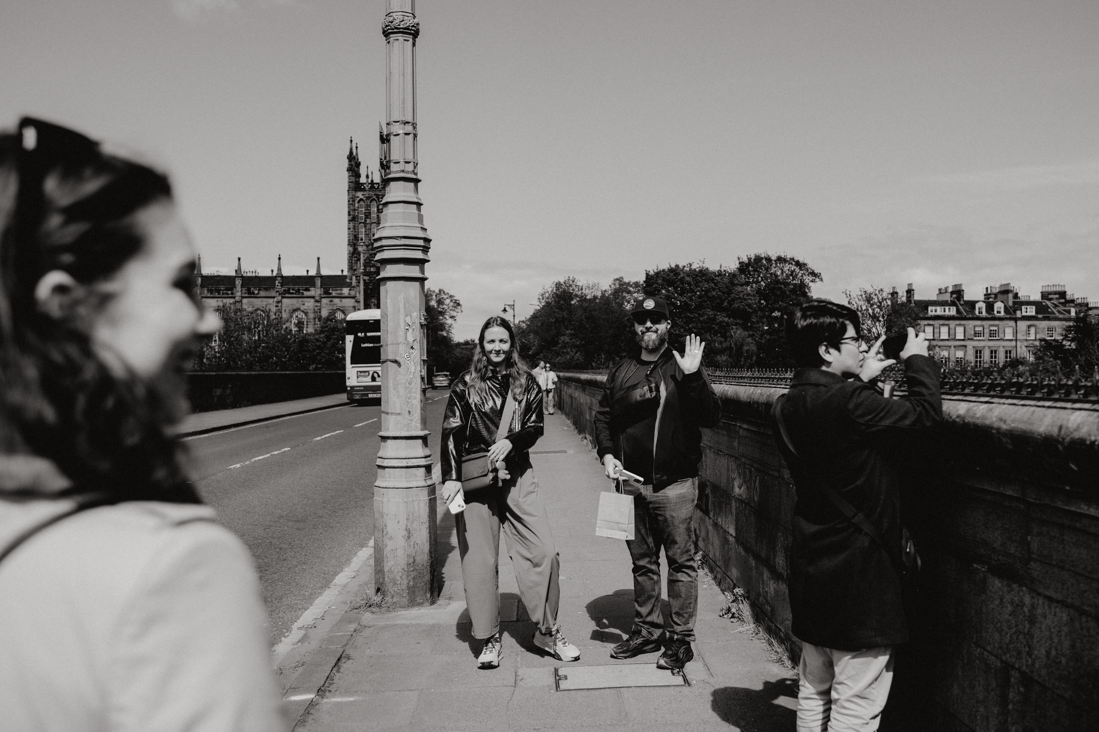 Two people stand on a bridge, one smiling and holding a phone, the other waving. In the background, a bus drives by and there are trees and buildings.