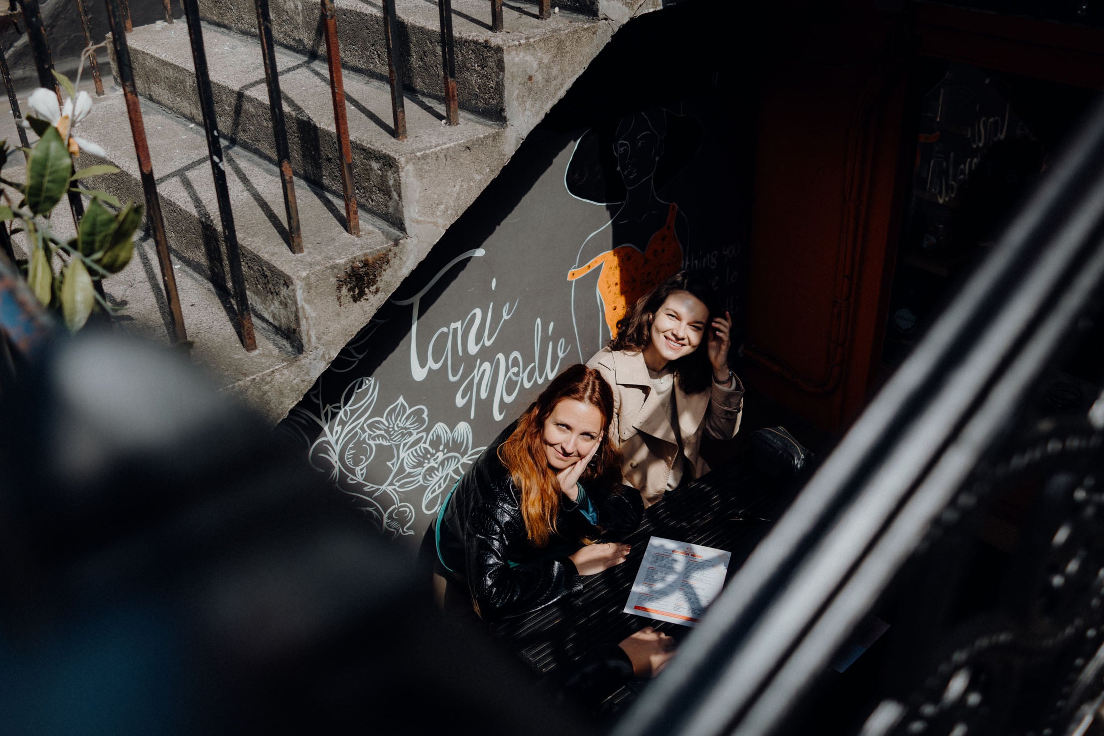 Two women sitting at a table in a cozy café, smiling and enjoying their time together. One woman has long red hair and is resting her chin on her hand, while the other has curly hair and is playing with her hair. The background features decorative wall art and a staircase.