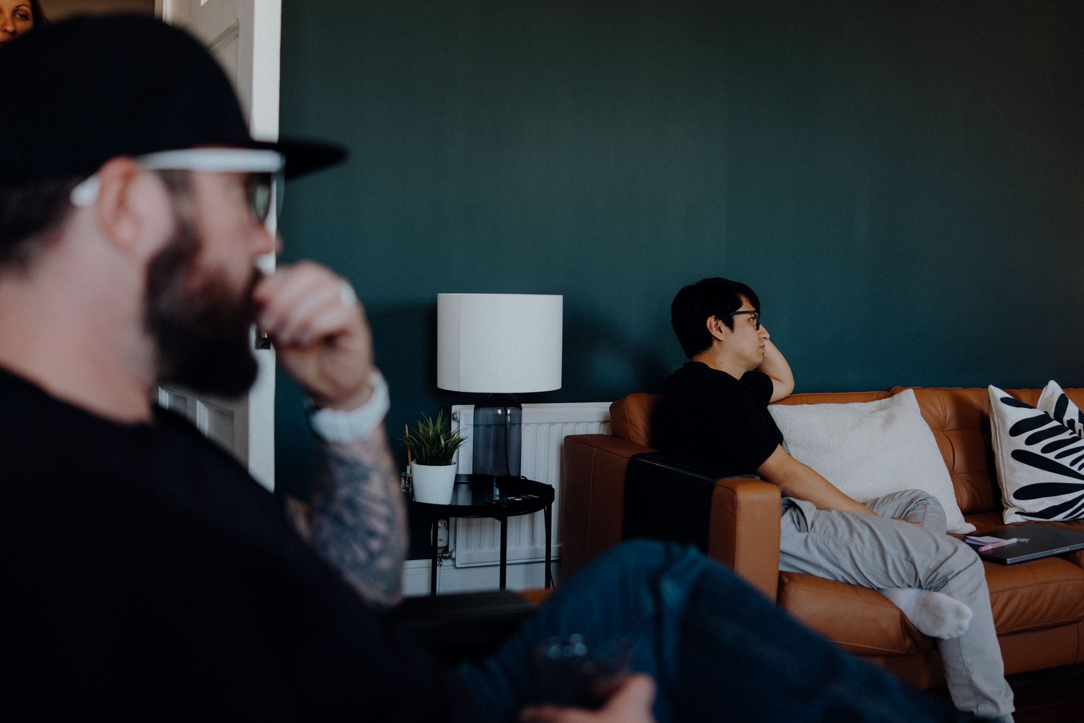 A man in a black shirt and baseball cap sits thoughtfully in a living room, while another man in a black t-shirt and glasses leans back on a brown leather sofa, looking contemplative. A lamp and small plant are visible in the background.