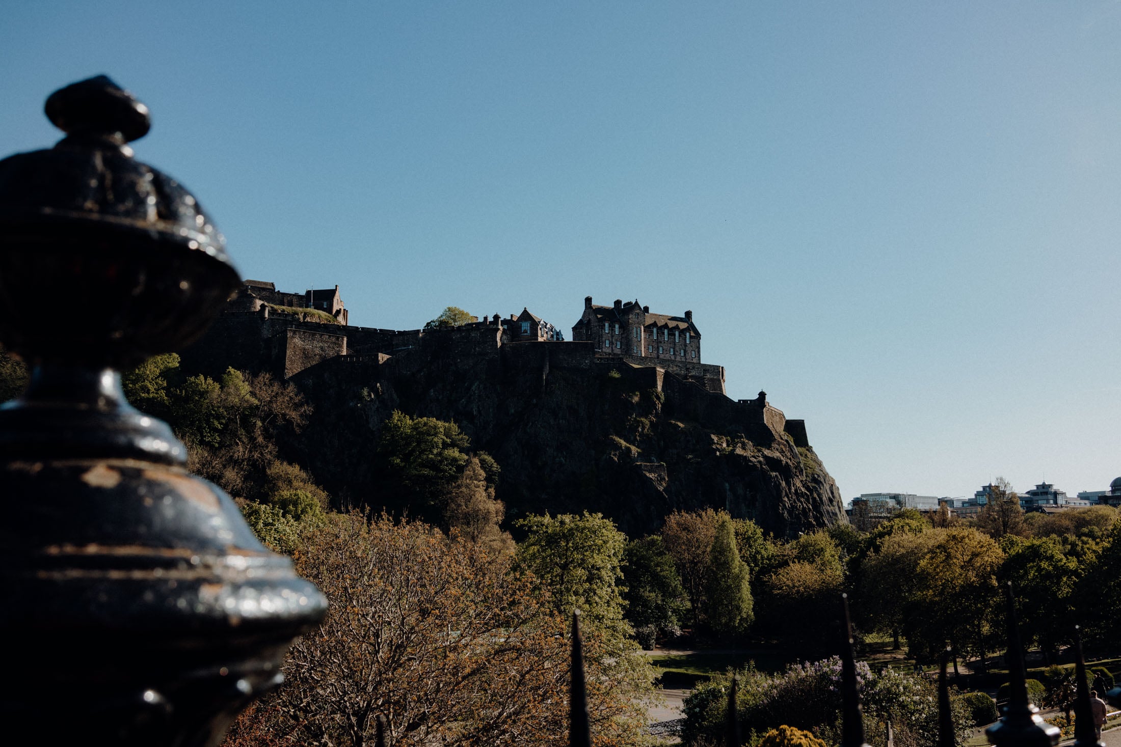 A view of Edinburgh Castle atop a rocky hill, with trees and a clear blue sky in the background.