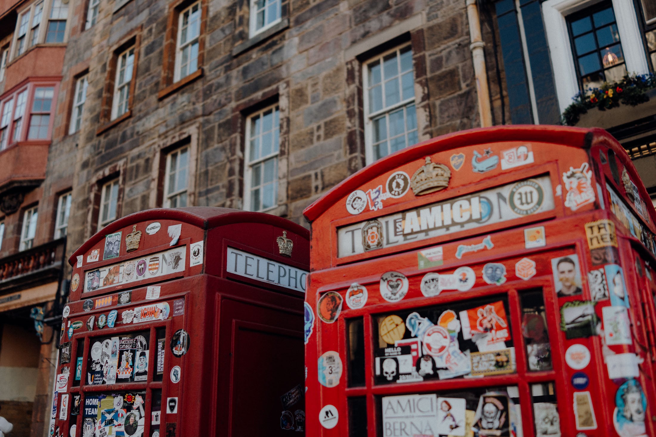 Two classic red British telephone boxes covered in various stickers, set against a backdrop of an old stone building with multiple windows.