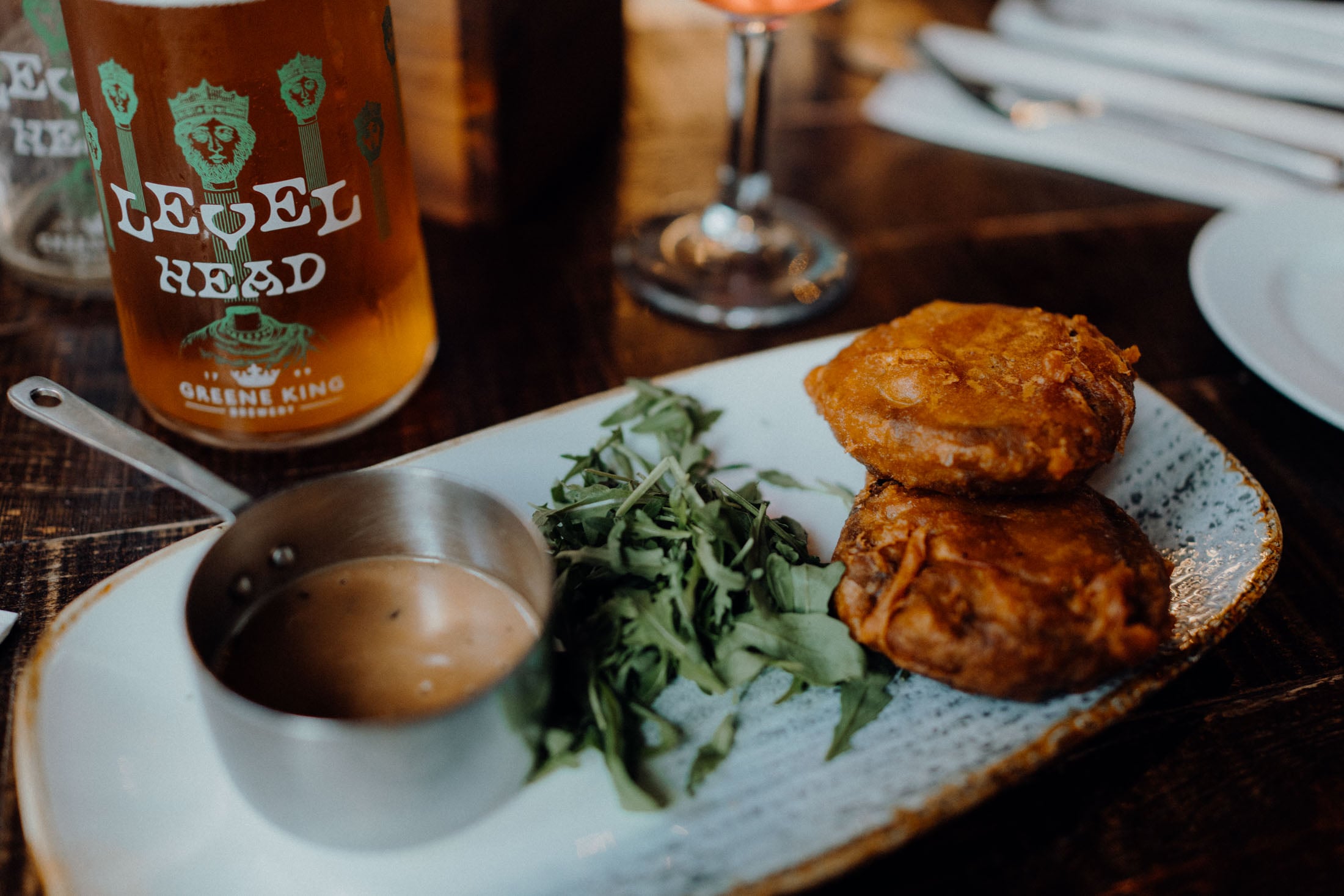 A plate with two golden-brown fritters, a small bowl of dipping sauce, and a serving of fresh greens, alongside a glass of Level Head beer.