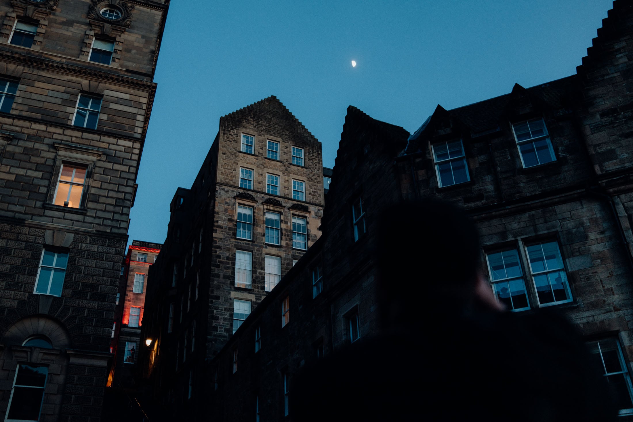 Silhouette of a person taking a photo with historic stone buildings and a crescent moon in the sky.
