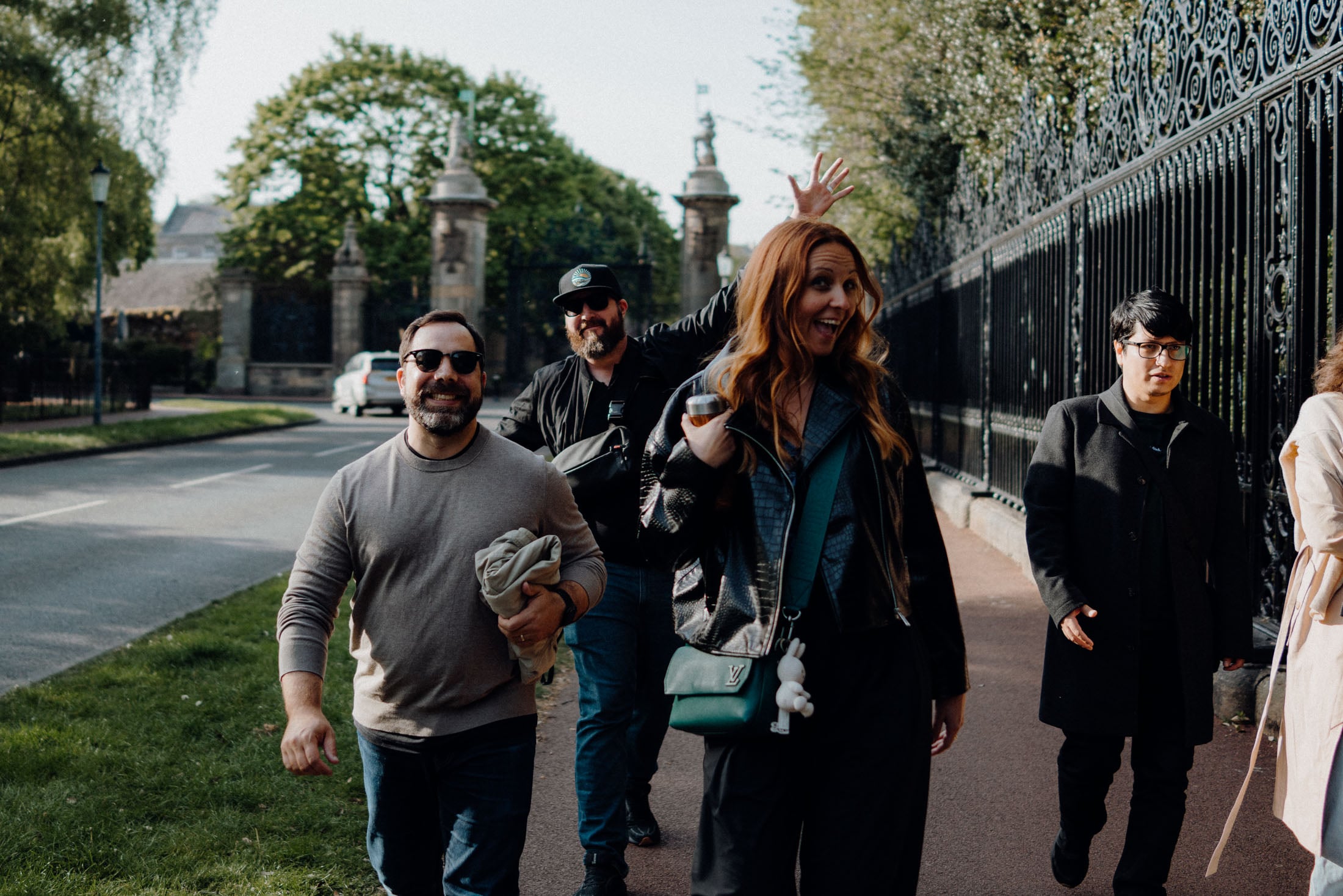 Four people walking on a tree-lined street, smiling and engaging with each other. The foreground shows a man in a beige sweater, a woman with long red hair wearing sunglasses, and another man with a beard wearing a black jacket. Another individual is seen in the background wearing glasses, walking beside them.