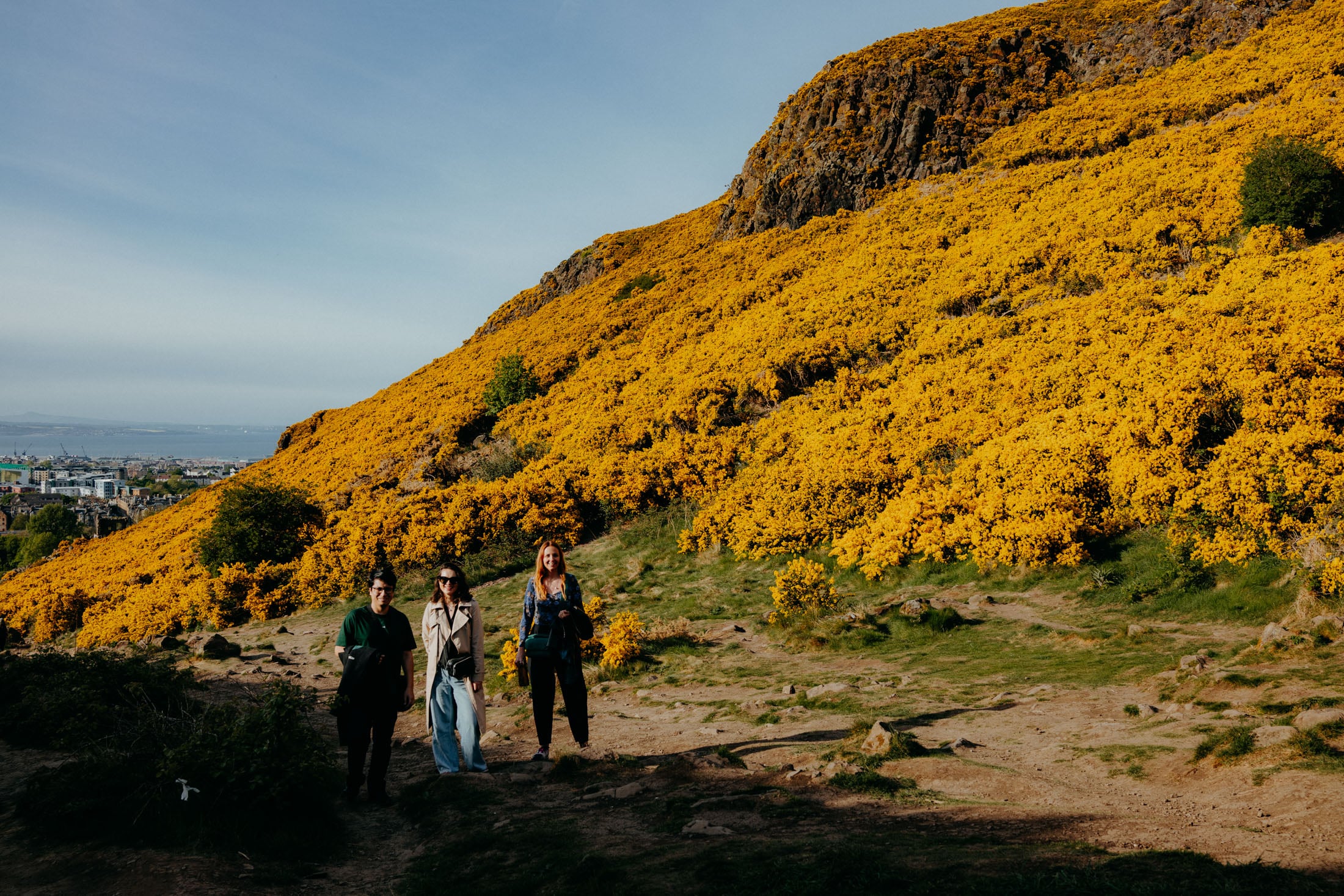 Three people walking on a path surrounded by vibrant yellow flowers and a scenic hillside in the background.