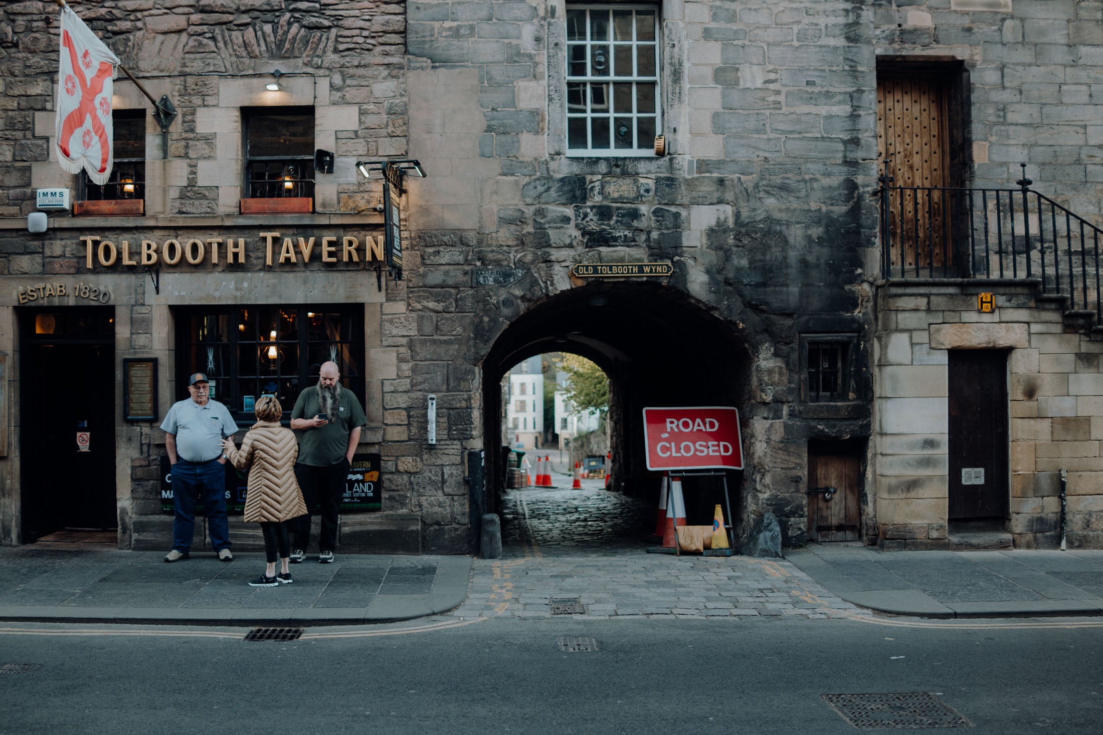 Exterior view of Tolbooth Tavern, featuring two men standing outside and a woman engaging with them, alongside a 'Road Closed' sign and an archway leading to another street.