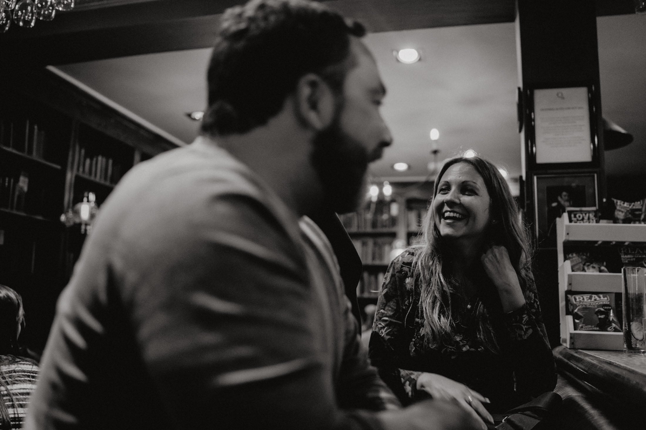 A man and a woman engaged in a joyful conversation in a cozy bar setting, with bookshelves in the background.