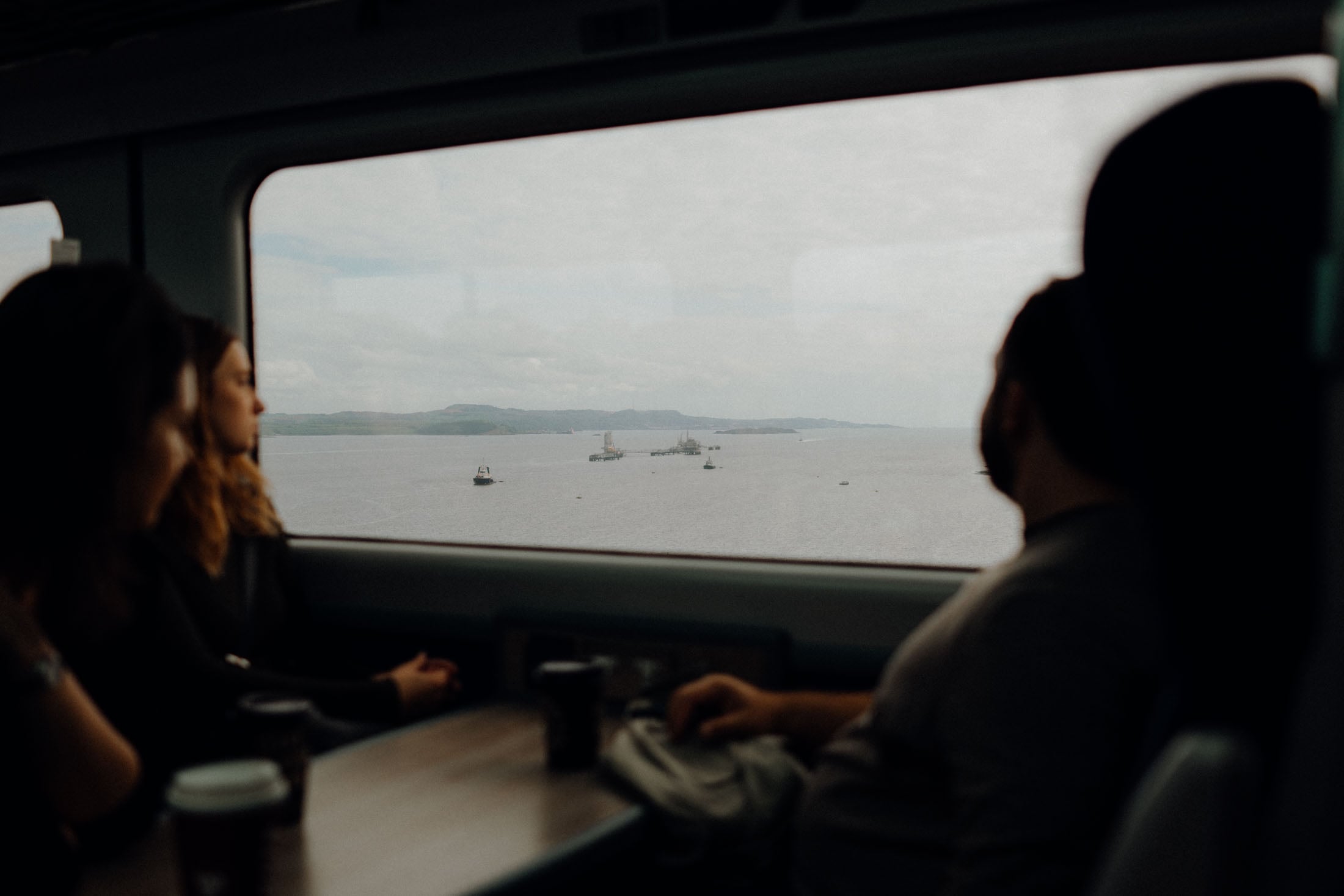 Two people sitting inside a train, looking out the window at a calm sea with small boats in the distance.
