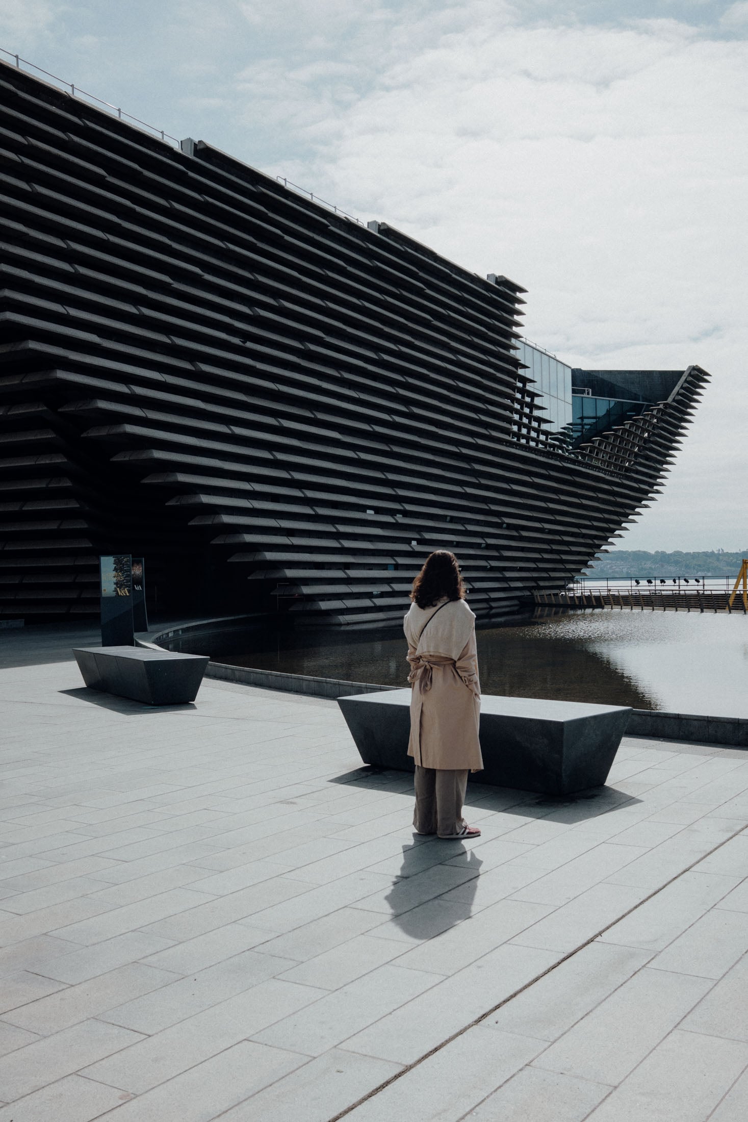 A woman in a beige trench coat standing in front of a modern architectural structure with layered design, beside a calm body of water.
