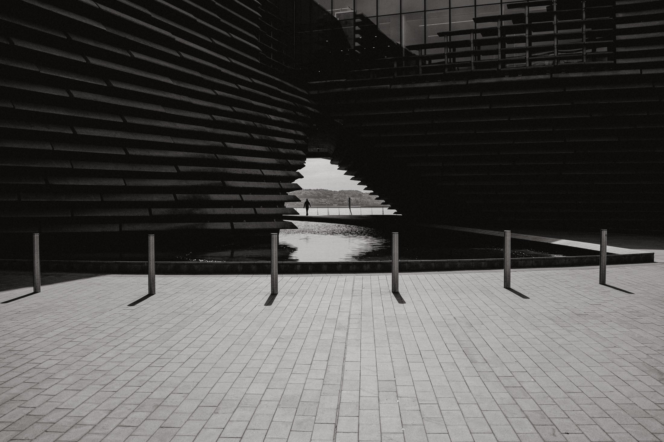 A modern architectural structure with layered, angular facades, framing a person walking towards a body of water in black and white.