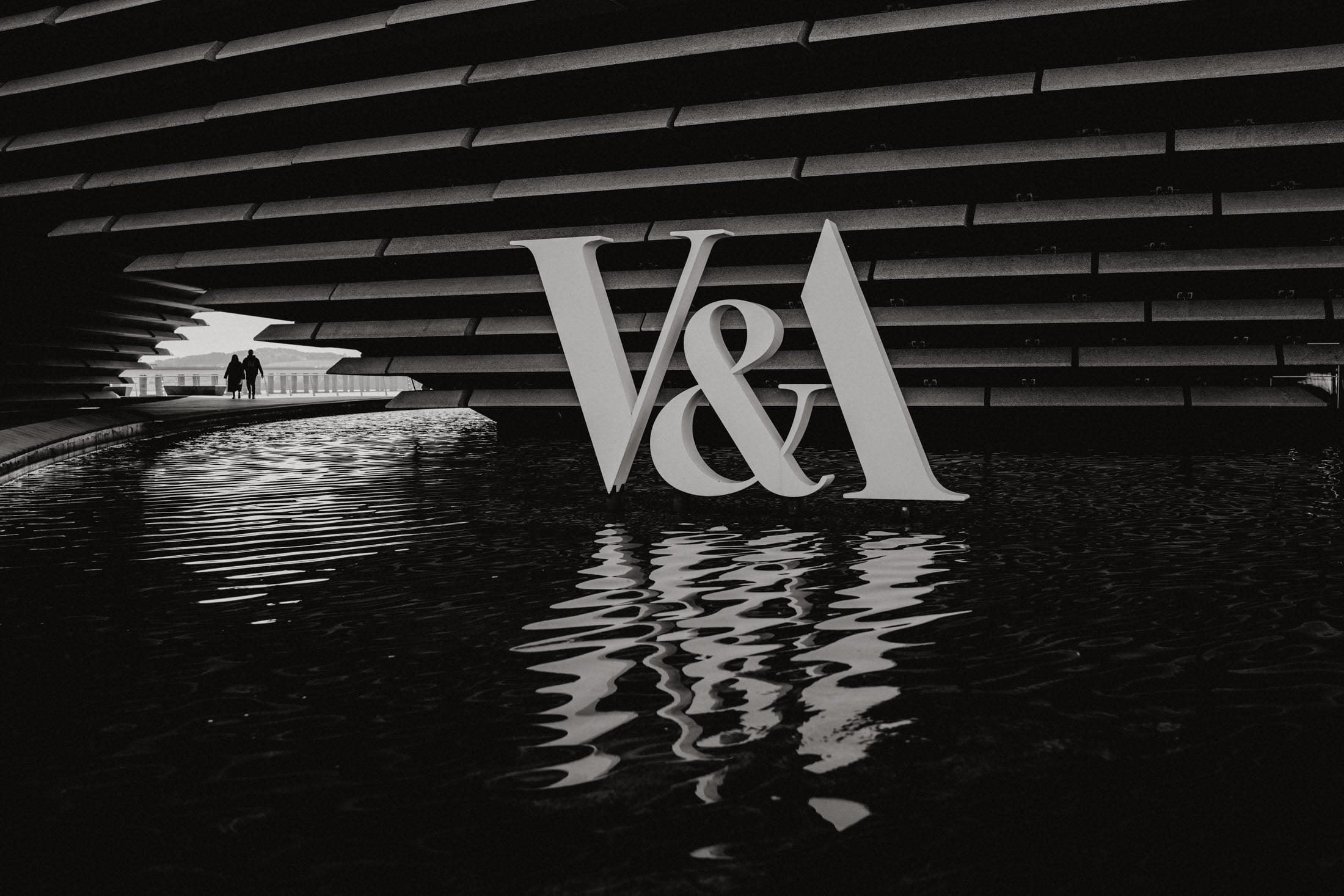 Black and white image of the V&A logo reflected in water, with two people walking in the background under a modern architectural structure.