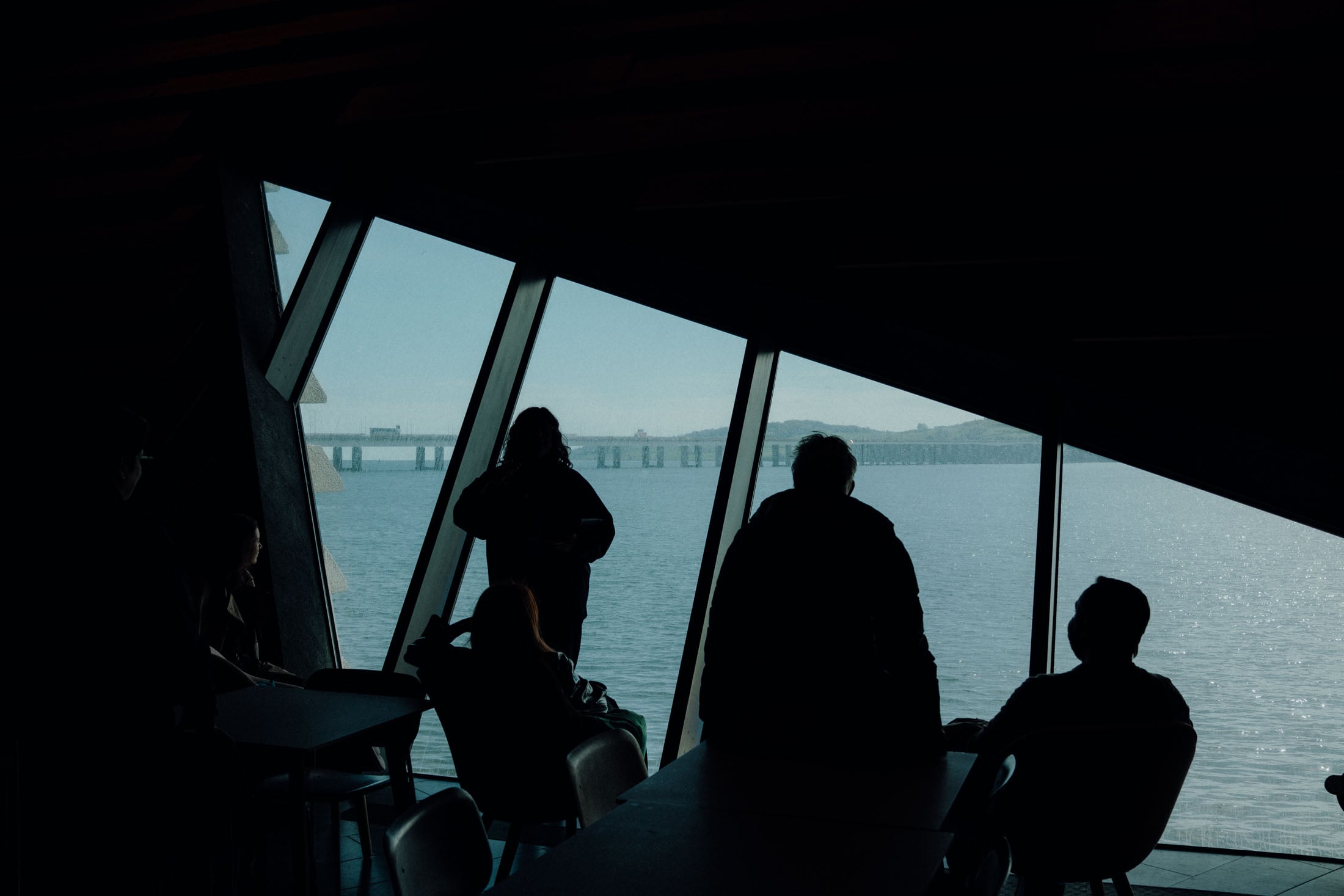 Silhouettes of people looking out a large, angled window towards a calm water view and a bridge in the distance.