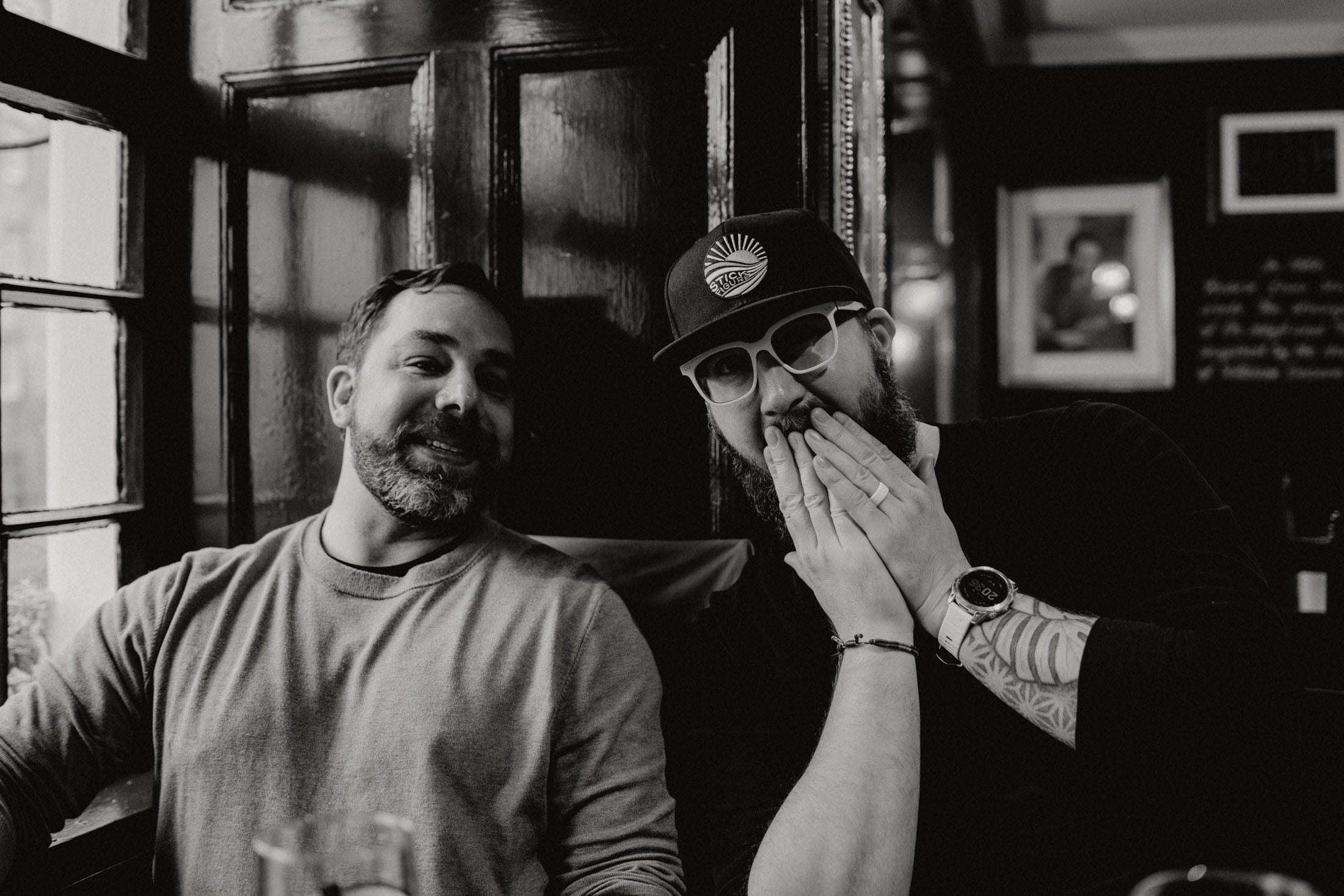 Two men sitting close together, smiling and posing for the camera, in a cozy, dimly lit space with wooden walls.