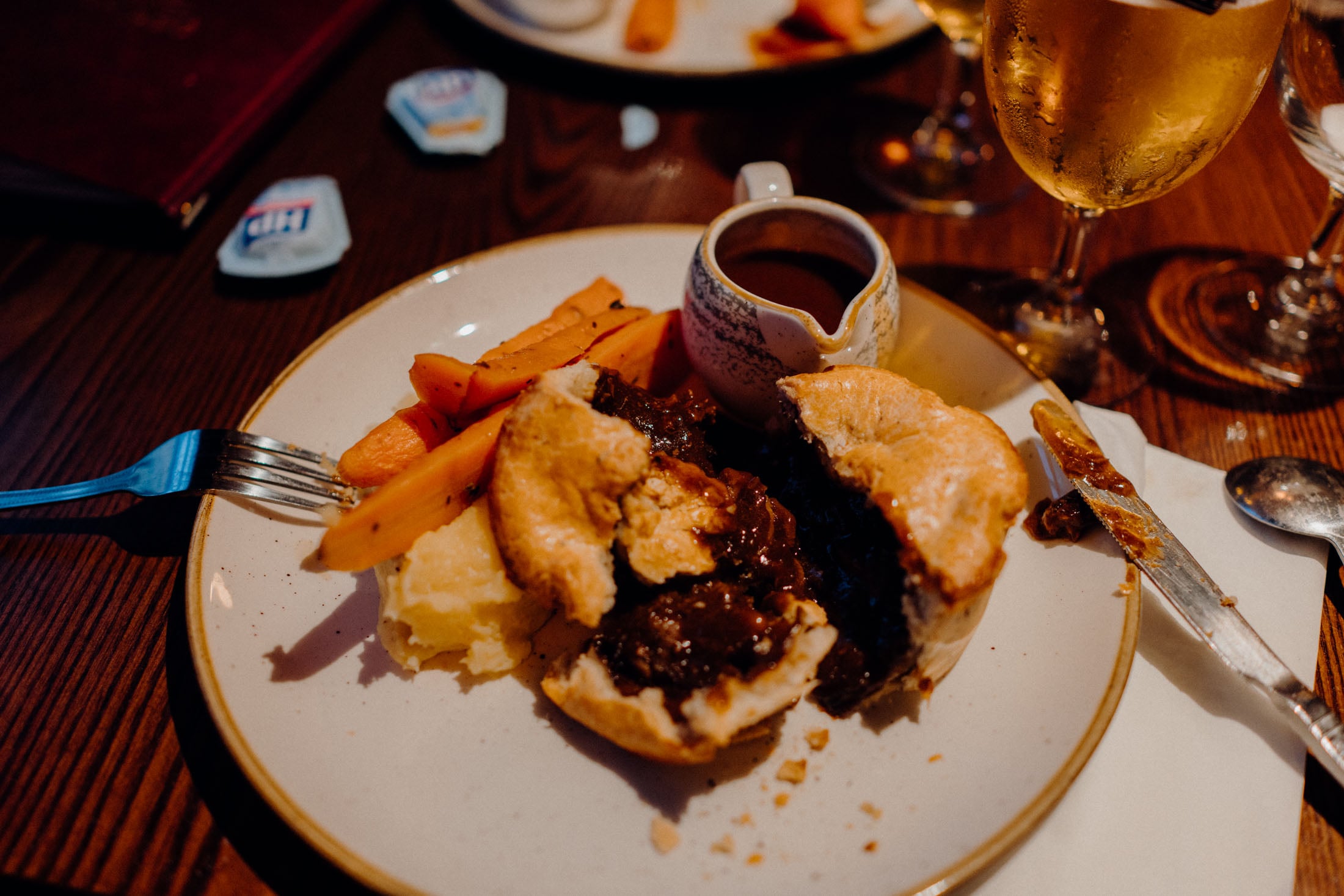 A plate featuring a meat pie filled with savory filling, accompanied by mashed potatoes and roasted carrots, along with a small jug of gravy and a fork.