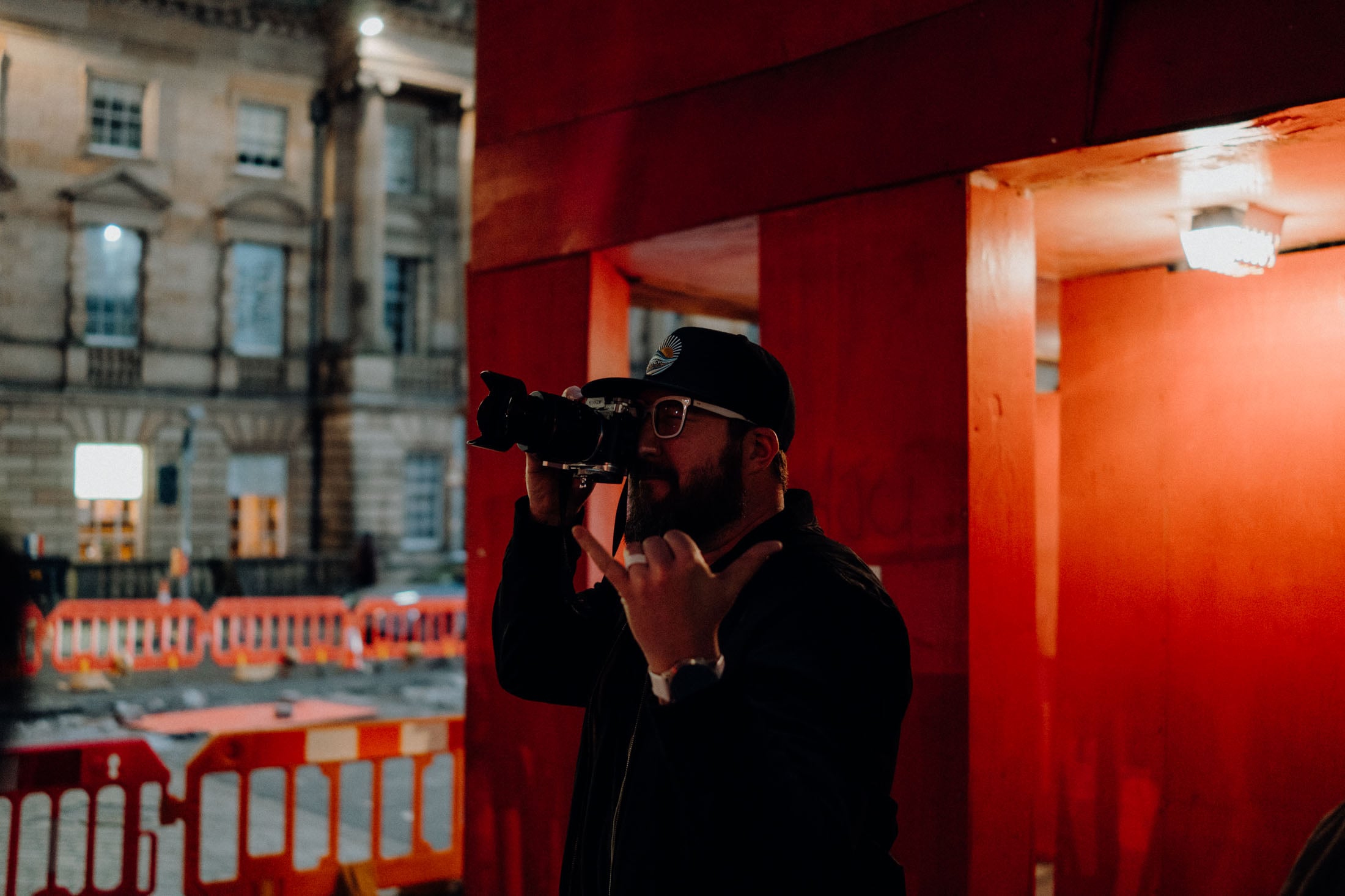A person wearing a black cap and glasses holds a camera, making a hand gesture, in front of a red wall.