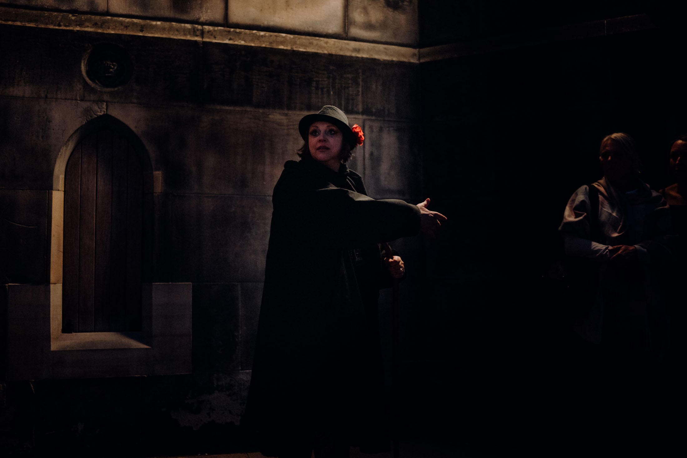 A performer wearing a black coat and a hat gestures while standing in a dimly lit space with stone walls, pointing towards a door.
