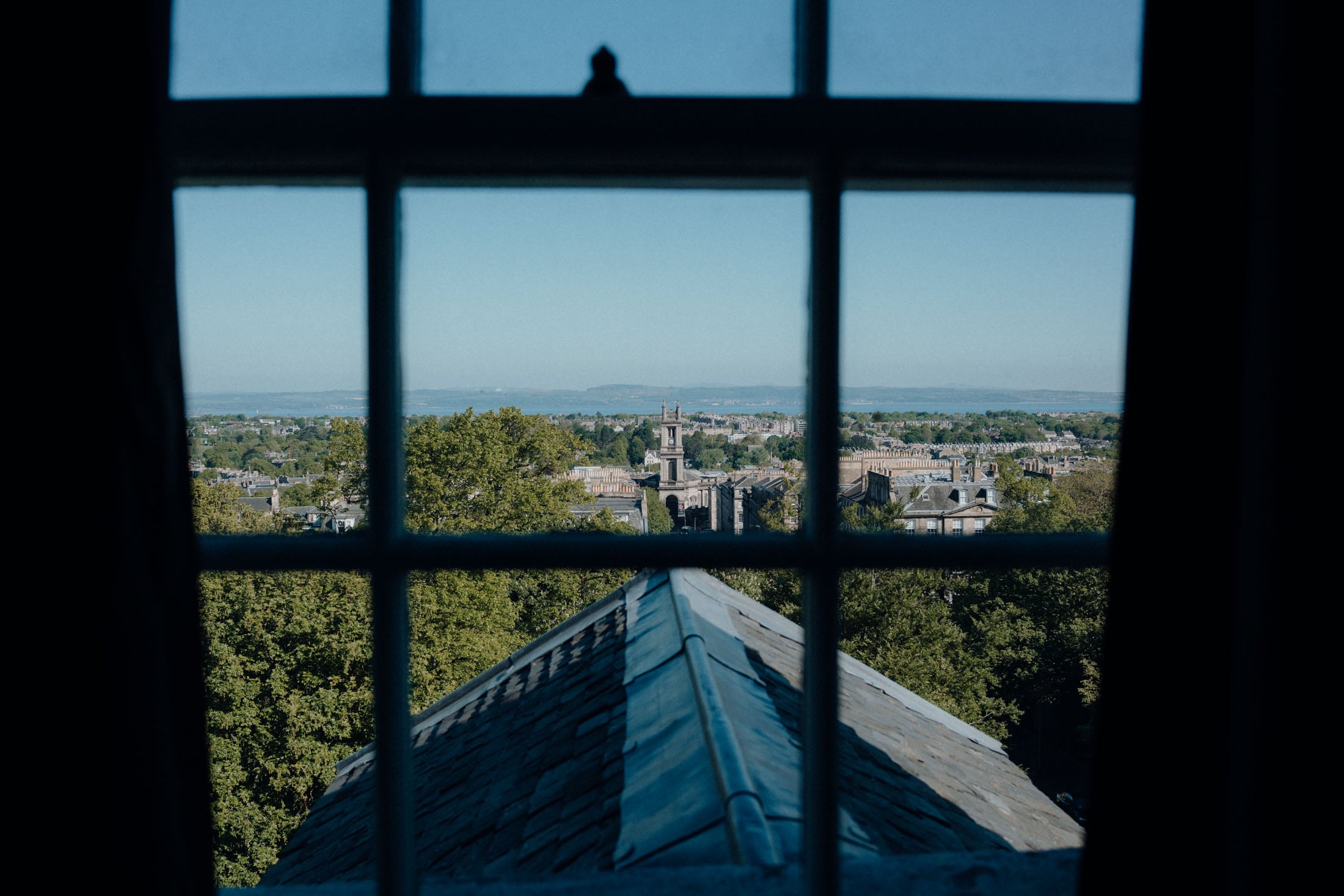 View from a window overlooking a cityscape with green trees, rooftops, and distant hills under a clear blue sky.