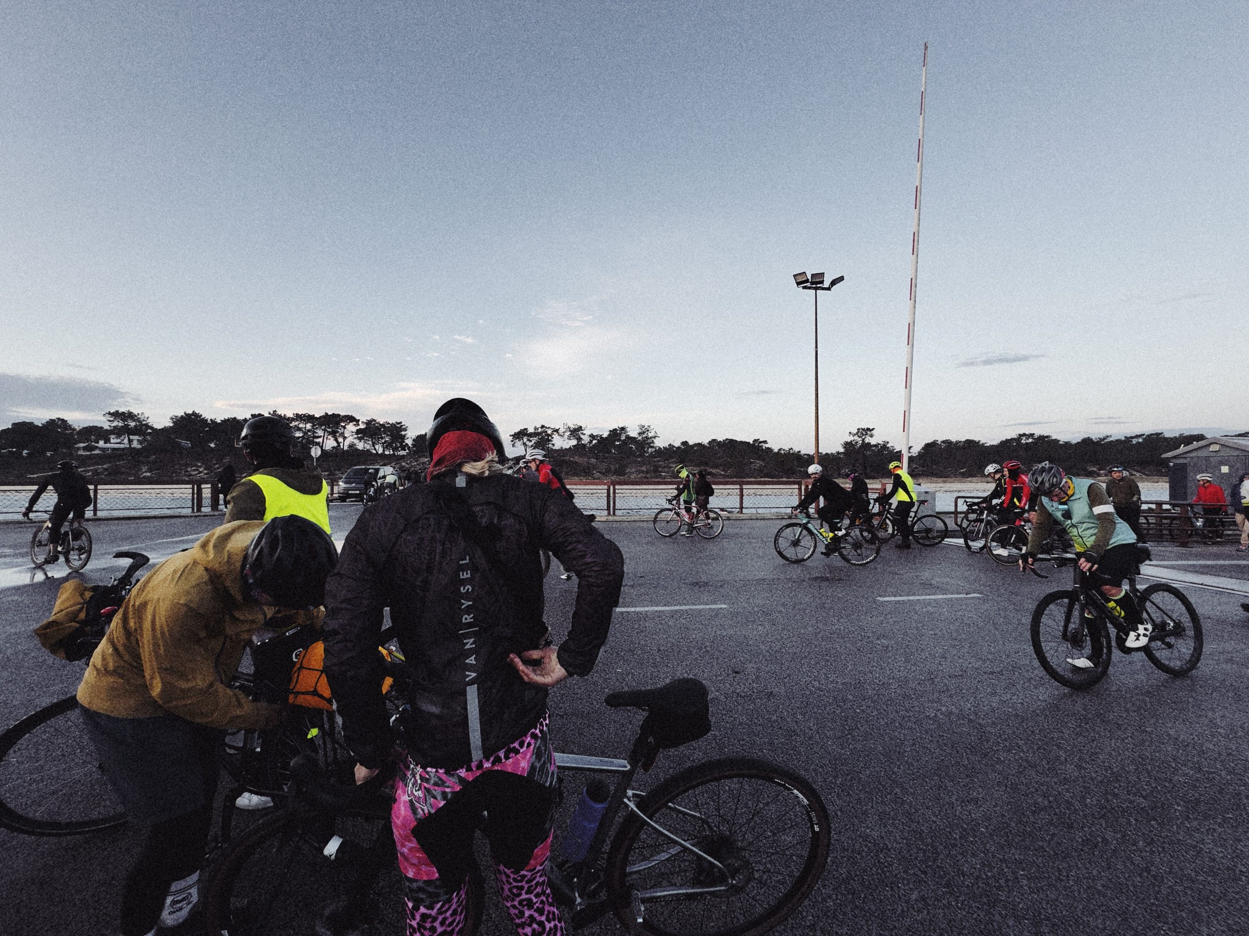 Cyclists preparing for a ride at dawn, some adjusting their bikes while others socialize in a group setting.