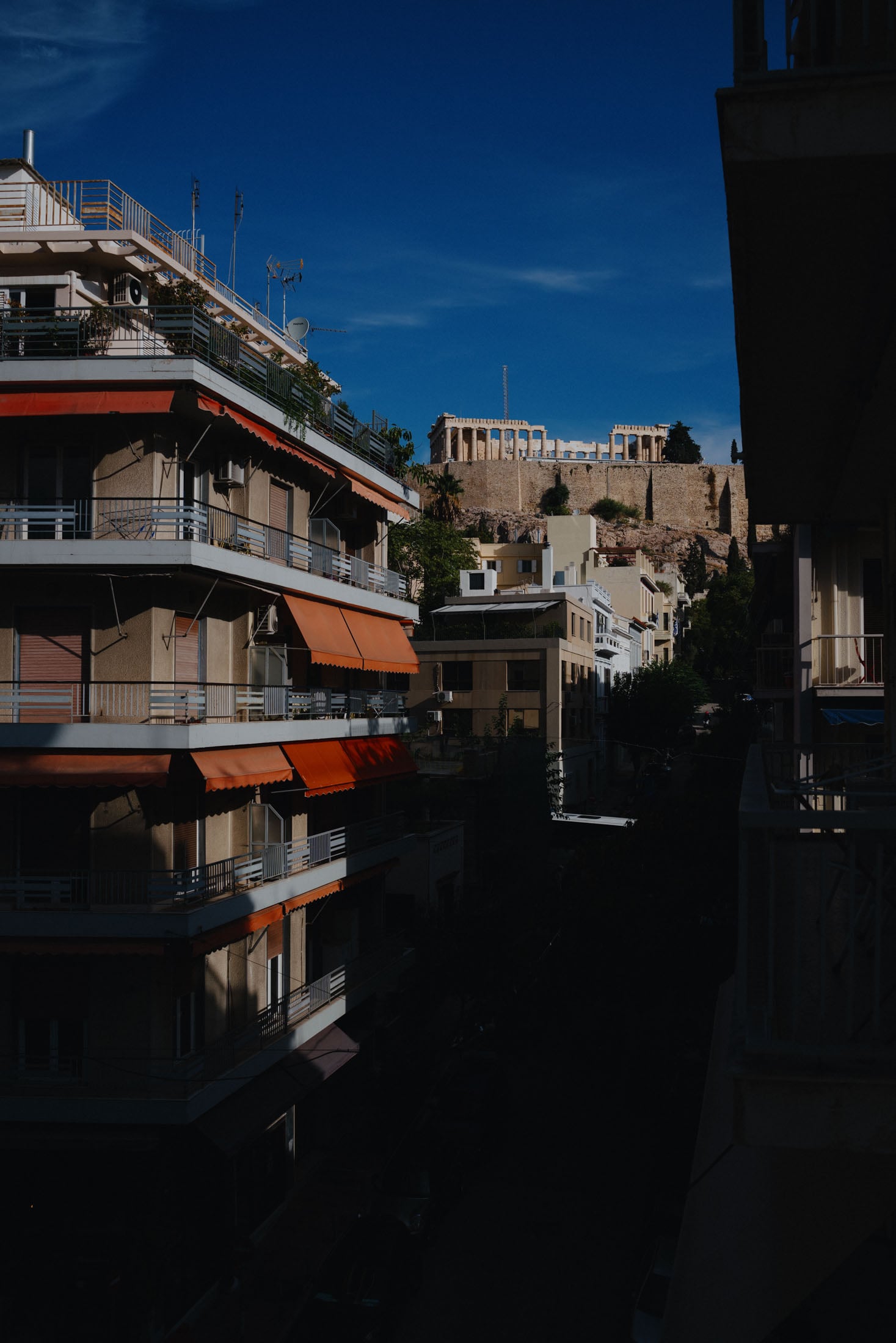 Cityscape view featuring buildings with orange awnings, set against a bright blue sky and the Acropolis in the background.