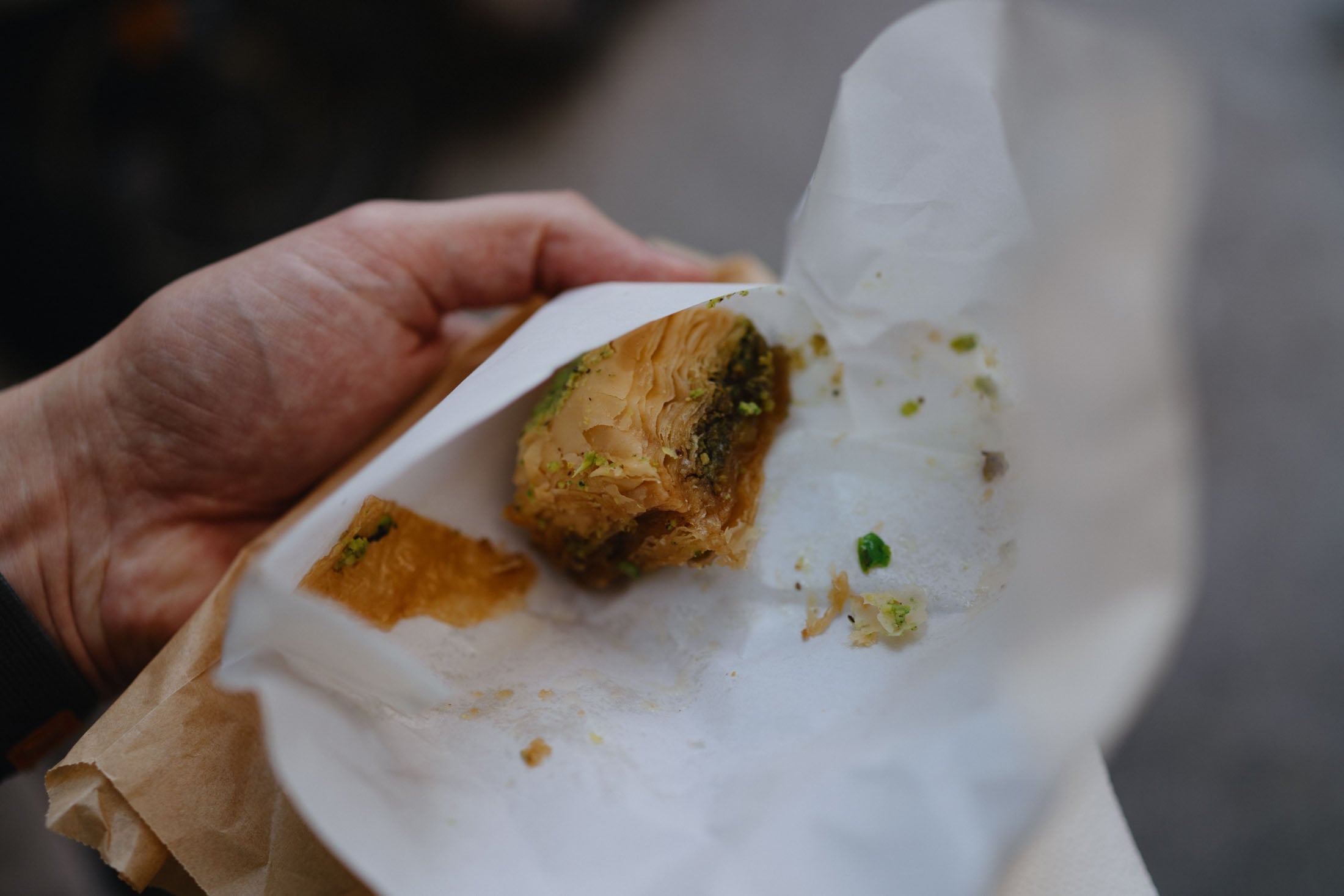 A hand holding a piece of baklava partially wrapped in parchment paper, showcasing layers of flaky pastry and chopped pistachios.