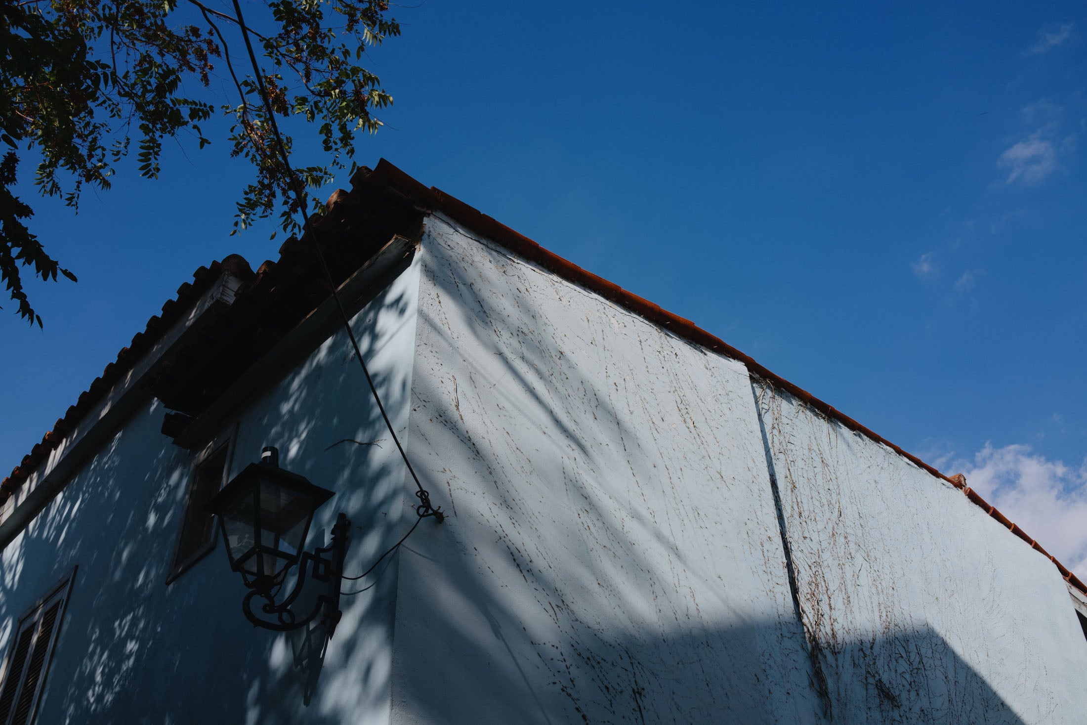Corner of a blue building with a tiled roof under a clear blue sky, featuring a vintage lamp and some shadows from nearby trees.