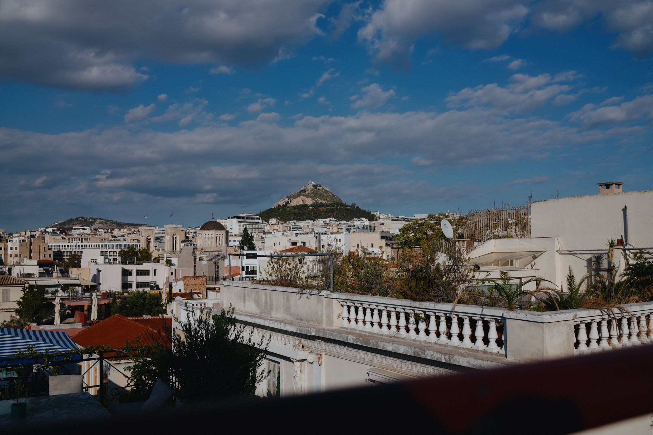 A panoramic view of a cityscape featuring buildings with a mix of architectural styles, a prominent green hill in the background topped with a structure, and a partly cloudy sky.
