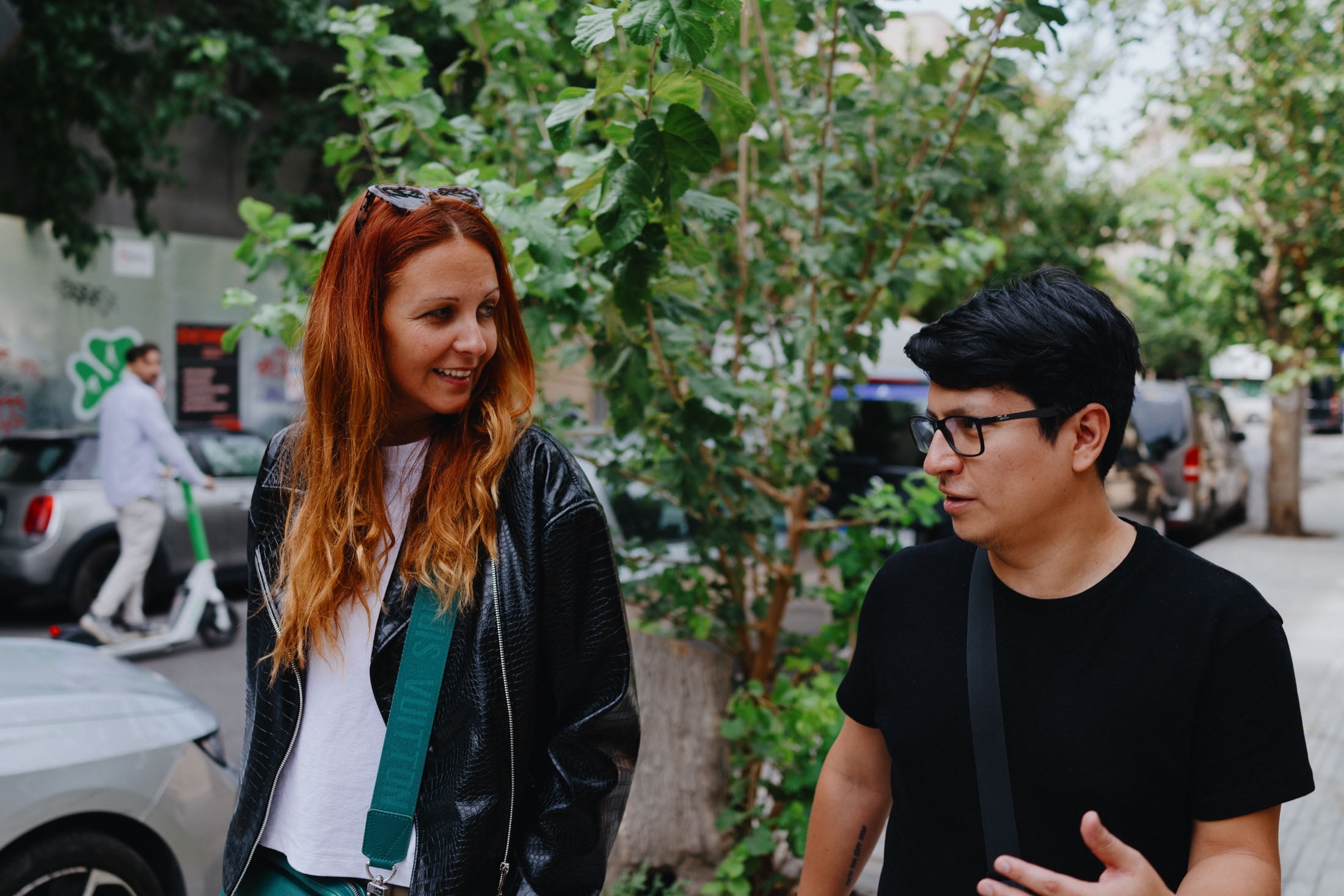 Two people walking and talking on a city street surrounded by greenery. The woman has long red hair, wearing a black leather jacket and a white shirt, while the man is wearing a black shirt and glasses.